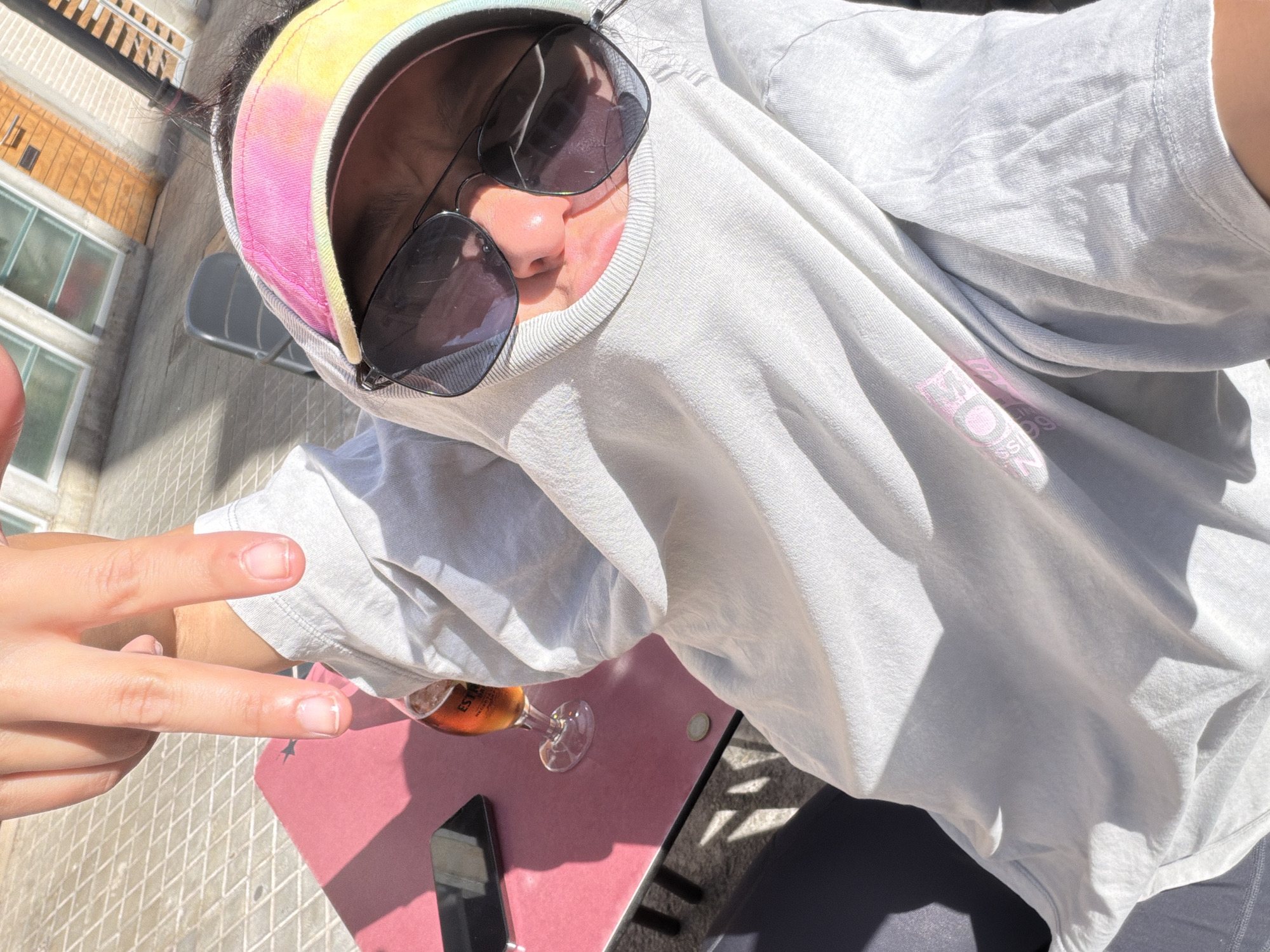 The image captures an outdoor scene, reportedly in the city of l'Hospitalet de Llobregat, Spain, under direct sunlight. A single individual is visible, positioned from a high-angle perspective. The person's head is enveloped by a light grey hooded sweatshirt. Beneath the hood, a visor displaying a pink, yellow, and light blue tie-dye pattern is worn. Dark sunglasses obscure the eyes and upper facial features, leaving only the lower portion of the nose and the mouth discernible. The person's left hand is raised, with the index and middle fingers extended to form a V-sign.
The individual is seated at a small, square, pink table. On the table, from left to right, a black smartphone is placed screen-down, a stemmed glass contains an amber-colored liquid with "ESTRELLA" branding visible on its side, and a small, round, bi-metallic object, consistent with a 1 euro coin, rests. Dark-colored leggings or trousers are visible on the person's lower body, below the table.
The ground surface consists of light-colored square tiles arranged in a diagonal pattern. Strong sunlight casts distinct shadows across the table and ground. In the background, a building with light-framed windows and a section of a horizontally paneled facade, possibly wood or brick, is visible. A grey metal chair is also present in the background.