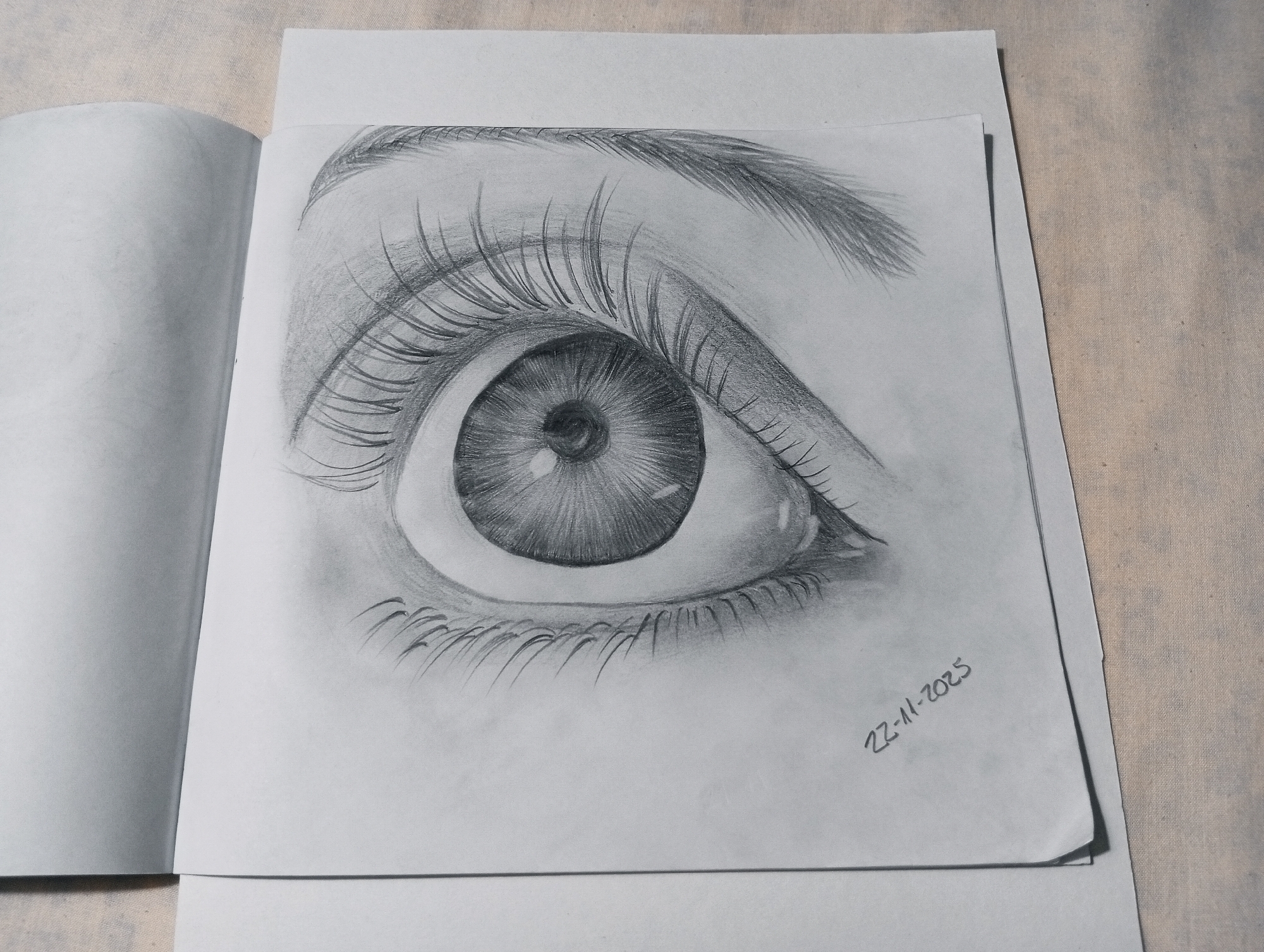 A close-up, overhead shot shows a graphite drawing of a human eye on a sheet of paper, resting on an open notebook. The drawing is detailed, capturing the iris, pupil, eyelashes, and eyebrow with varying shades of graphite to create depth and realism. The eye is wide open and appears to be looking slightly to the viewer's left. Below the drawing, in the bottom right corner of the paper, is a handwritten date in graphite: "22-11-2025". The paper is placed on top of a textured, light-colored fabric surface, possibly a tablecloth or backdrop. The left side of the image features the slightly unfocused edge of an open notebook, with blank pages visible. The drawing itself appears to be on a separate sheet of paper that is slightly curved, with its bottom edge overlapping another piece of paper or the notebook. The overall lighting suggests a soft, ambient light source from the right, casting subtle shadows. The scene is static and appears to be an artist's work-in-progress or a finished piece displayed on a table. The image has no discernible connection to San Antonio de Los Altos, Venezuela.