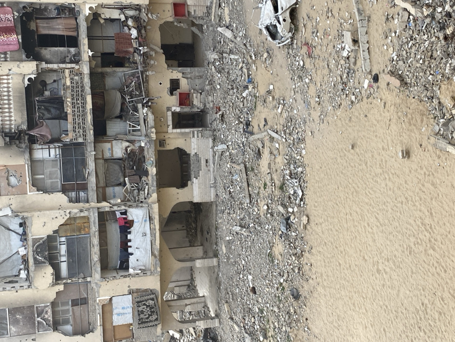 An overhead view captures a heavily damaged urban scene in Khan Yunis, Palestinian Territory. A multi-story residential building dominates the background, exhibiting extensive structural damage, numerous impact holes consistent with bullet marks, and widespread destruction of windows, balconies, and exterior walls. Many interior spaces are exposed, revealing debris and scattered belongings; clothes are visibly hanging on a line from a central balcony, and a patterned rug is draped over a lower-left balcony railing. UNICEF-branded materials are visible in some window openings. The ground floor features damaged arched entryways. The area directly in front of the building is covered with a dense layer of rubble, including concrete fragments, metal debris, and various remnants of destroyed structures. To the right, the skeletal remains of a white vehicle, stripped of most body panels, rest amidst the debris. The lower-left foreground consists of a large, flat expanse of light brown sand, sparsely dotted with smaller pieces of rubble. No individuals are present in the frame.