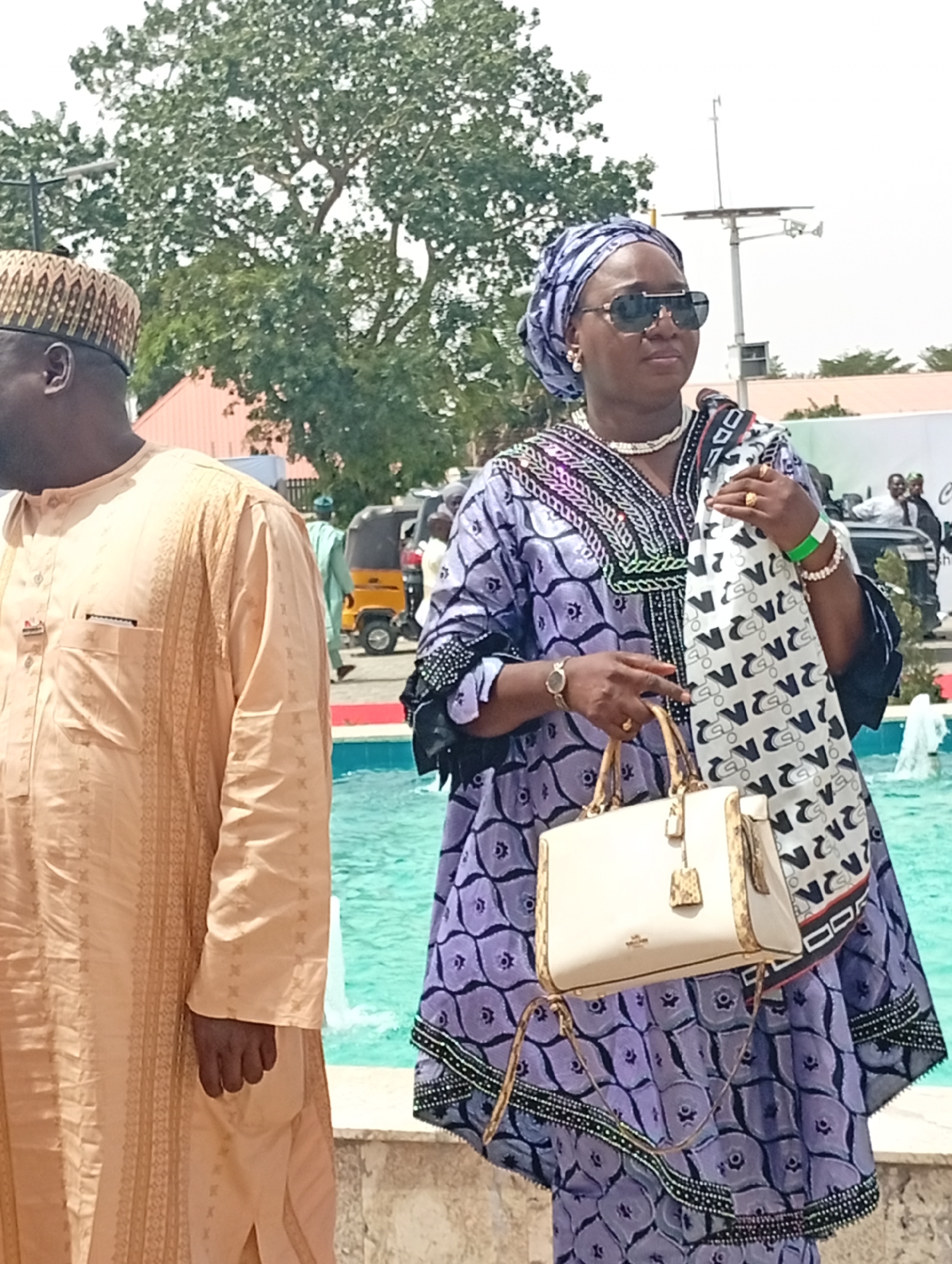 An outdoor scene in Unguwan Sanusi, Nigeria, depicts two individuals in traditional attire by a body of water, likely a fountain or pool. The bright daylight suggests a sunny day.
On the right, a woman stands prominently, facing slightly away from the camera. She wears a striking long-sleeved traditional dress in purple with black patterns and embroidery, complemented by a matching headwrap. Her accessories include large dark sunglasses, a white beaded necklace, white pearl earrings, a gold watch, a gold ring, and white beaded bracelets. A green wristband is visible on her right wrist. She holds a cream-colored handbag with gold hardware and has a white scarf with black, stylized patterns draped over her left arm. Recognizable text on the scarf includes "ZARA" and repetitive letter-like designs, possibly "N" or similar glyphs.
To her left, partially visible, stands a man in a light beige or cream-colored traditional outfit, possibly an agbada, featuring subtle vertical patterns. He wears a patterned, ornate cap. His right hand is visible by his side.
In the background, a light blue fountain or pool adds a vibrant element to the scene. Beyond this, scattered trees and faint outlines of buildings are visible. Further back, vehicles, including a yellow auto-rickshaw (keke napep), and other people can be seen, suggesting a public gathering or event. A tall pole with cameras or antennae is also in the distance. The overall impression is that of a social occasion in a public space.