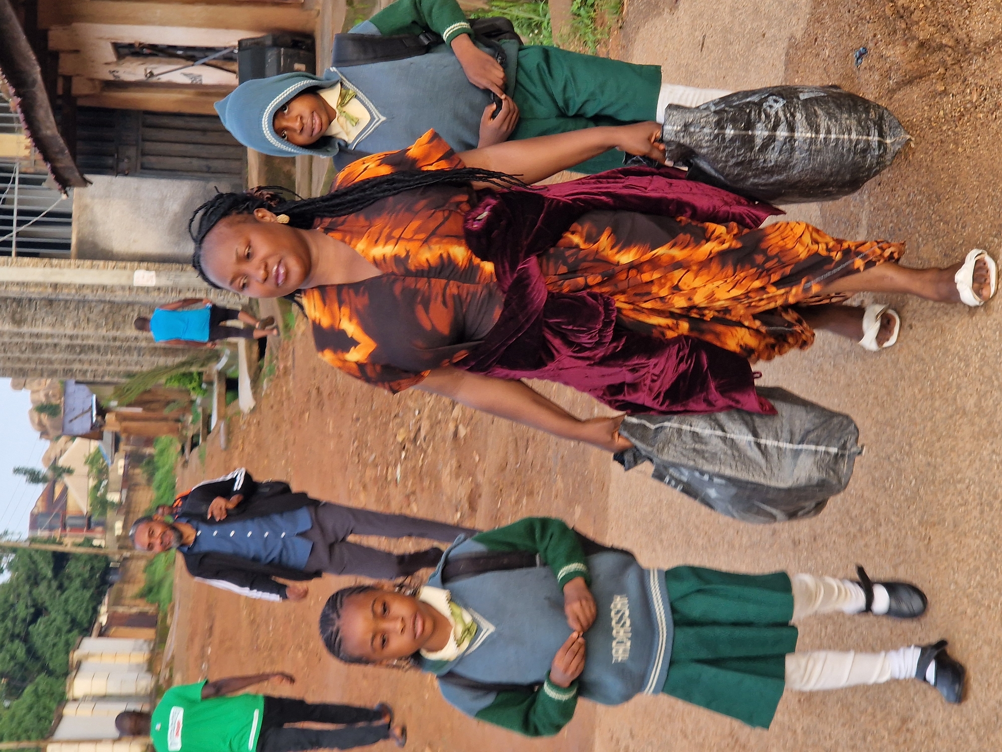 In Kabong, Nigeria, on a dirt road lined with low-rise buildings and scattered vegetation, a group of people walk together. A man in a green shirt walks away from the camera, followed by a man in a black jacket and blue shirt. Closer to the foreground, a woman in a vibrant orange and black patterned dress walks with a large black plastic bag. Beside her, a young person wearing a blue hooded sweatshirt with a tie and a grey sweater vest, also carries a black plastic bag. In front of them, a young girl in a green skirt and a blue and green sweater vest walks with her hands clasped. The environment suggests a residential or commercial area in a developing urban setting.