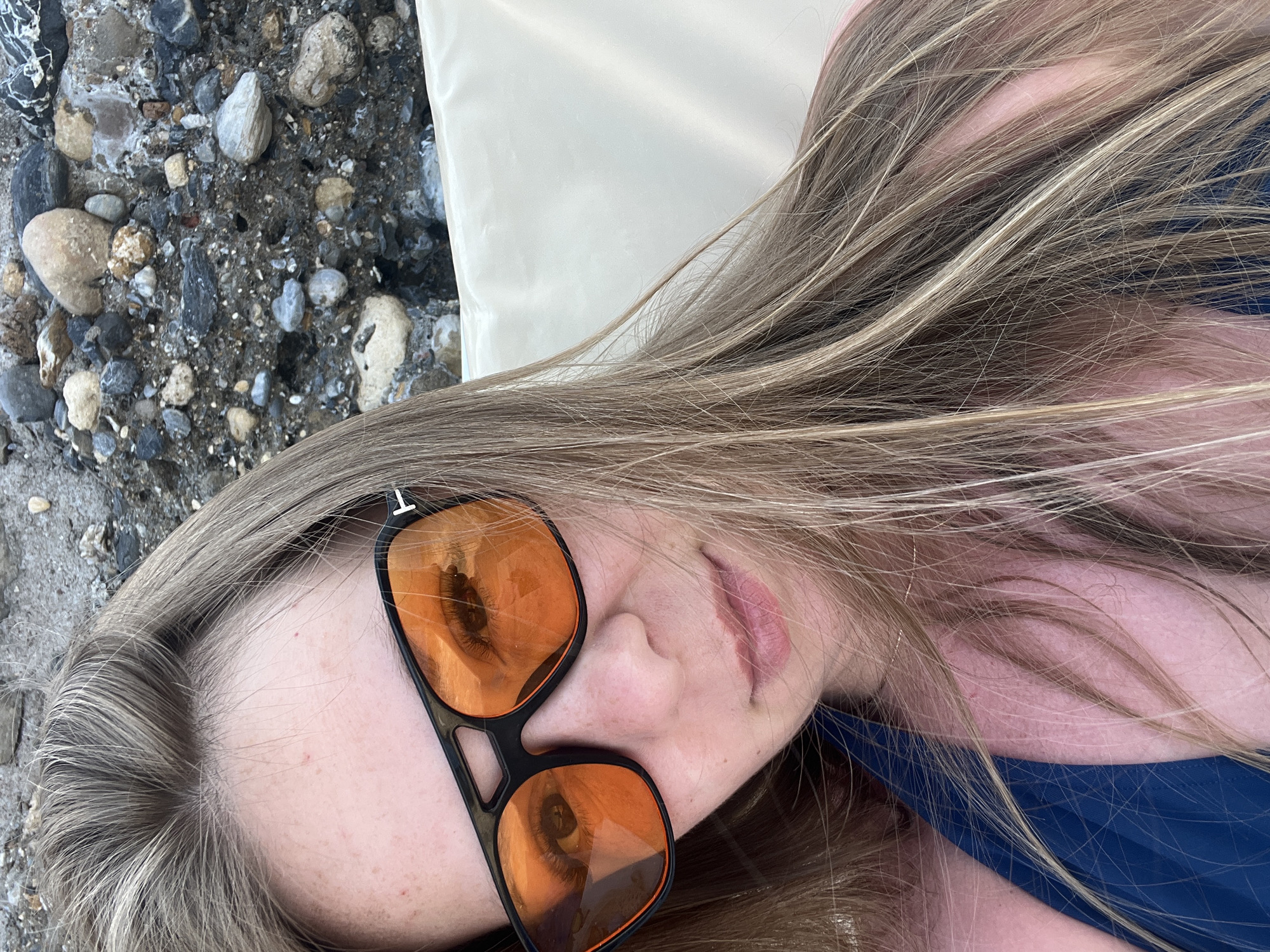 A woman, viewed from the chest up, lies on a light-colored lounger. She wears a blue swimsuit and black sunglasses with orange lenses. Her blonde hair is partially obscuring her face and shoulders. The background consists of a rocky, pebbly surface, likely on a beach. The overall setting suggests a relaxed, outdoor environment in Alanya, Turkey.