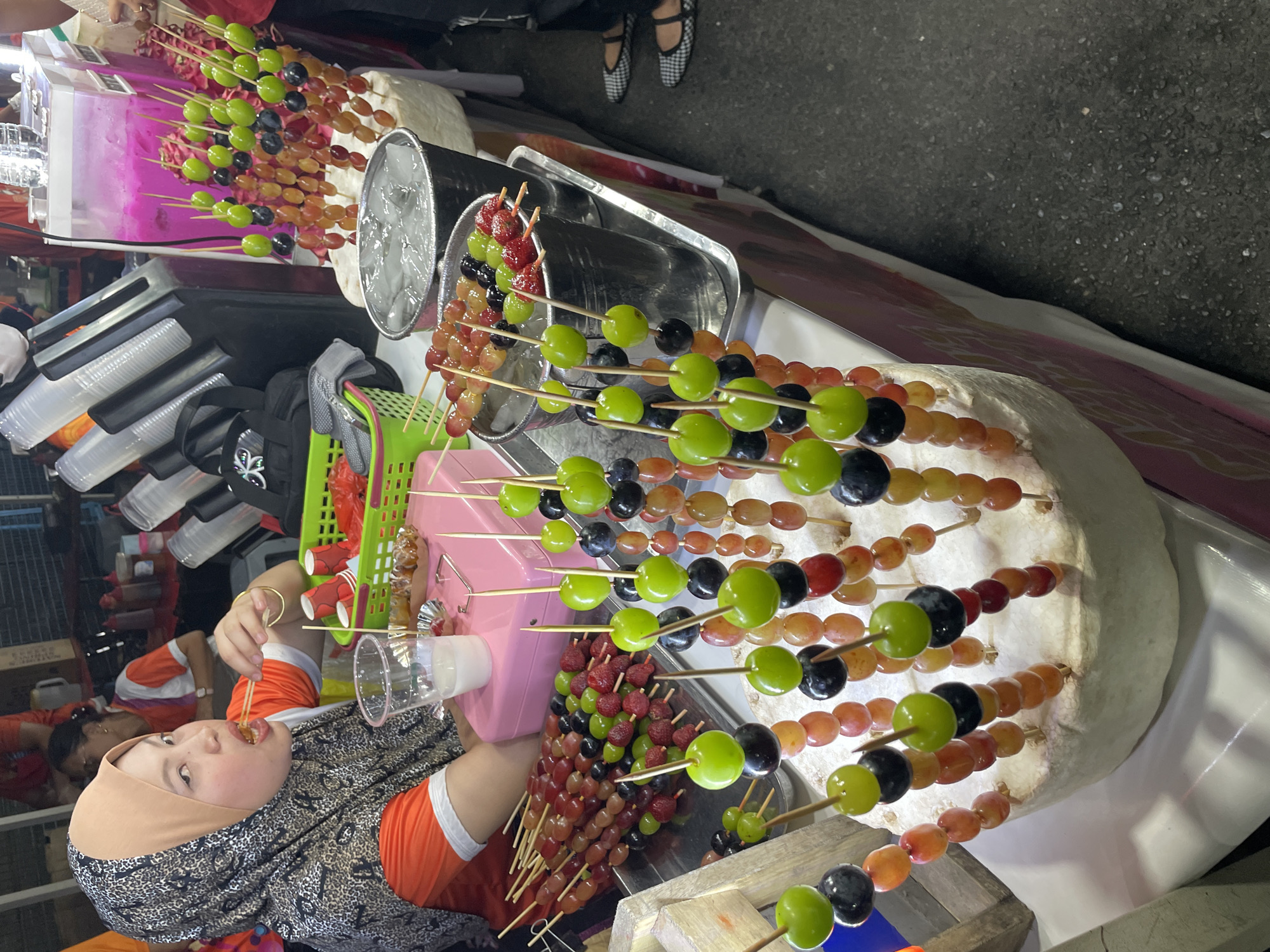 The image depicts a street food stall, likely located in Davao City, Philippines, specializing in candied fruits. A woman wearing an orange shirt and a patterned hijab is visible in the foreground, actively eating a skewer of candied fruits. She is holding a skewer with her right hand and has her mouth open as she bites into the fruit. To her left, a clear plastic cup containing a milky white beverage is placed on a pink tray, along with another skewer of candied fruits.
The stall displays an assortment of candied fruits on wooden skewers, arranged vertically on a cylindrical white base. These fruits include green grapes, red grapes, black grapes, and strawberries, all coated in a glossy, hardened sugar glaze. Some skewers are placed in metal buckets filled with ice. In the background, there are stacks of clear plastic cups, a pink-hued beverage dispenser with what appears to be dragon fruit pieces, and other food items or supplies. The ground surface is asphalt, typical of an outdoor market or street setting. The overall scene suggests a vibrant and busy street food environment.