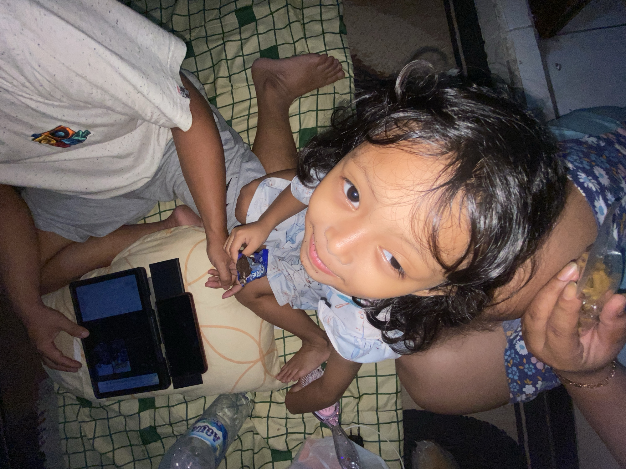 The media file shows a close-up, overhead view of a young child and other people in what appears to be a home setting in Gelam, Indonesia. The child, with dark, curly hair, is looking up at the camera with a curious expression. They are wearing a light blue shirt with a subtle pattern.
In the foreground, there are several objects laid out on a checkered mat. These include a tablet displaying images, a smartphone, and a partially unwrapped chocolate snack that the child is holding. To the left, a partially visible person is wearing a white t-shirt with a colorful graphic on the chest and grey shorts. Another person's legs and arm are visible on the right side of the frame, and this person is wearing a blue floral patterned garment. A bottle of Aqua brand mineral water is also present.
The lighting suggests it might be evening or indoors with artificial light. The overall atmosphere seems relaxed and informal, possibly indicating a family gathering or downtime. The focus is primarily on the child's engagement and the surrounding items. No specific event is explicitly depicted, but the presence of the tablet and snacks suggests a casual activity.