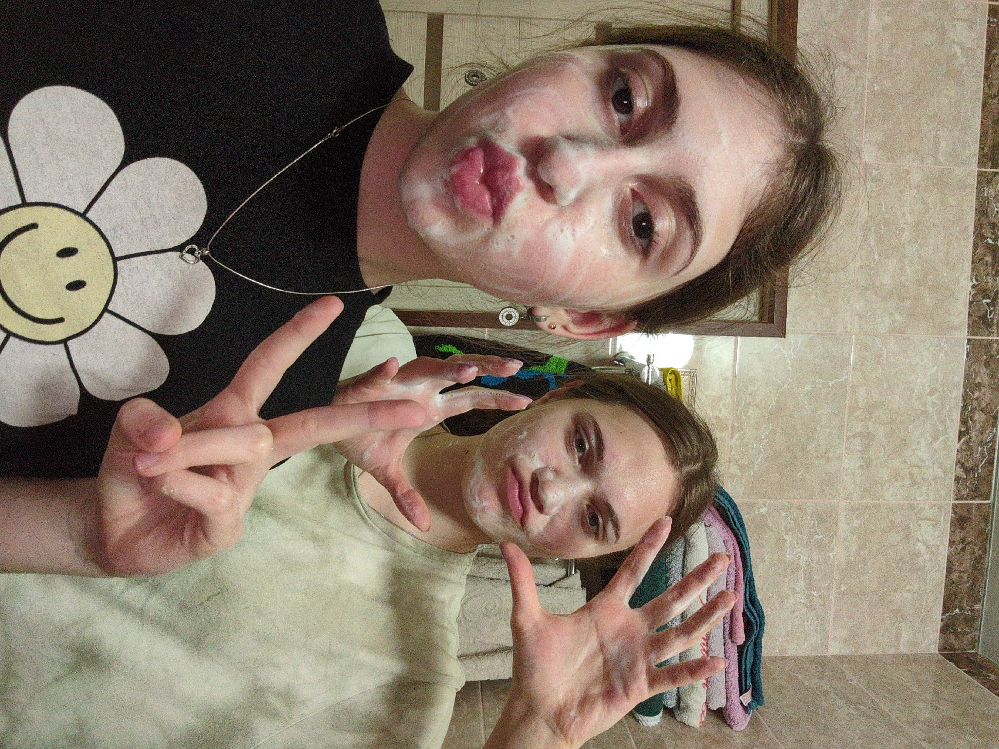 Two young women are posing for a selfie-style photograph in a bathroom with light-colored tiled walls. Both have a white soapy substance on their faces, presumably a face mask. The woman at the top of the frame is wearing a black t-shirt with a large white daisy graphic and a silver necklace with a heart-shaped pendant. She is making a kissing face towards the camera. The woman at the bottom is wearing a light-colored t-shirt. She is holding up her hands, fingers spread wide, towards the camera, and she is also smiling slightly. A stack of colorful towels is visible behind her. A framed mirror is partially visible behind the top woman. The location is Unknown, Unknown.