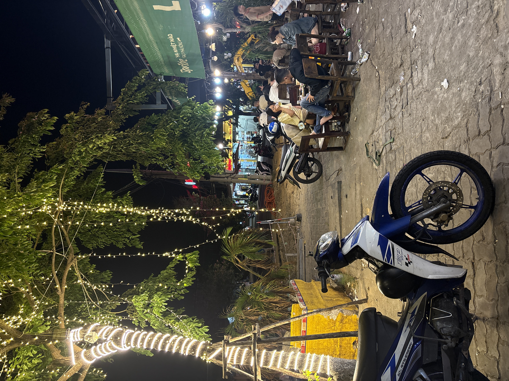 The scene shows an outdoor dining area at night in Đà Nẵng, Vietnam. Several people are seated at wooden chairs and tables along a sidewalk, likely a restaurant or street food vendor. The area is illuminated with string lights hanging from trees and overhead structures. A motorcycle is parked in the foreground, and other motorcycles are parked further down the street. The street surface is paved with brick or stone. A green sign with Vietnamese text is visible above the seating area. Construction or repair work seems to be happening nearby, as indicated by a yellow barrier.