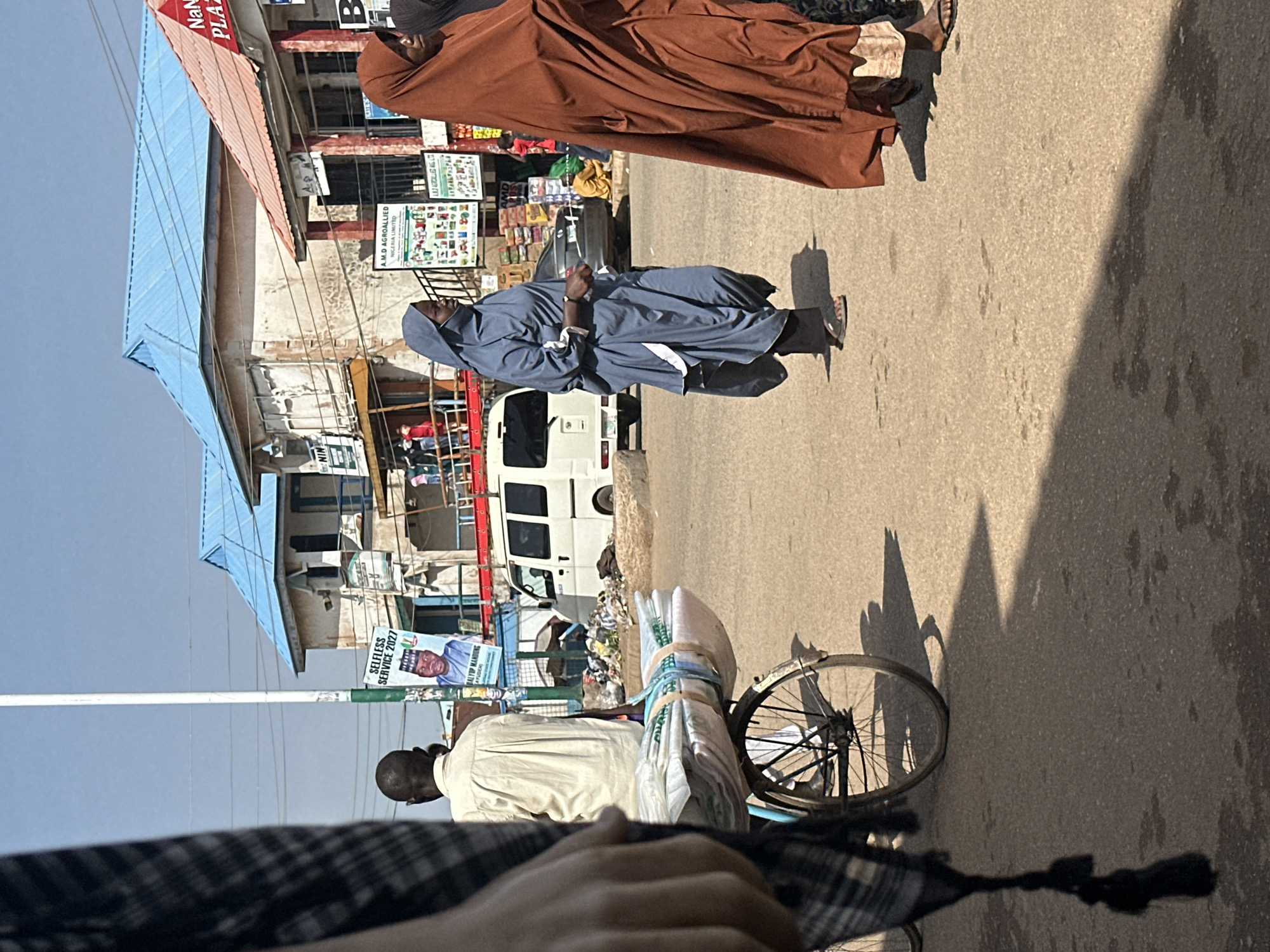 In the city of Bukuru, Nigeria, a street scene unfolds under a clear sky. A man in a cream-colored shirt is positioned behind a bicycle laden with rolled-up white material secured with blue and green straps. His back is to the camera, and he is looking towards a pole with a poster featuring a political candidate.
To his right, a woman wearing a blue hijab and abaya walks across the dusty road. Her abaya has white trim, and she is wearing dark pants and sandals. Further to the right, another woman in a rust-colored hijab and abaya is partially visible, also wearing sandals.
The background shows buildings with blue and orange-red roofs. One building has a sign that reads "Nanai Plaza." Another sign displays "A.M.D AGROALLIED NIGERIA LIMITED." A white van is parked on the side of the road, and stacks of goods are visible in front of some of the shops. Debris is scattered on the ground near the van. The ground is paved and appears to be asphalt, with shadows cast by the people and objects, indicating a sunny day. A dark, checkered fabric hangs in the foreground on the left side of the frame.