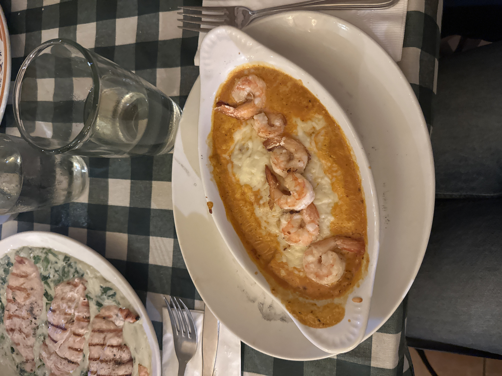 A top-down shot shows a green and white checkered tablecloth on a table. Two dishes and two glasses of water are visible. The first dish, on the right, is a white, oblong ceramic dish filled with a reddish-orange sauce, covered with melted cheese and topped with several shrimp. This dish rests on a larger white plate. A fork and knife are placed beside the plate. The second dish, located on the lower left side, contains grilled chicken breasts with a creamy sauce and spinach. A second set of silverware is located next to the dish. The location is implied to be Naucalpan de Juárez, Mexico.
