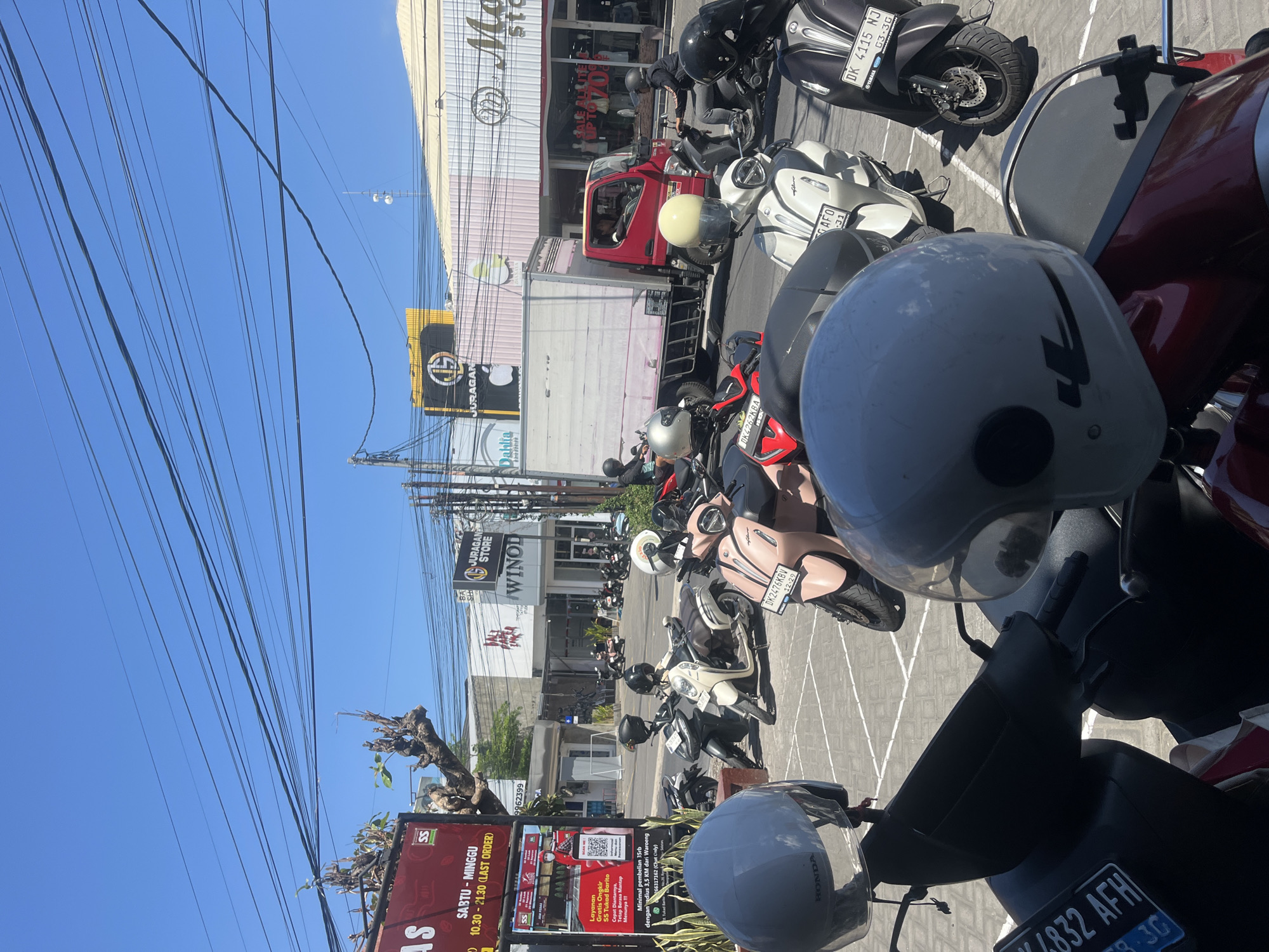 The image captures a street scene in Denpasar, Indonesia, on a sunny day. The foreground is dominated by several parked scooters and helmets, with a red scooter in the bottom right partially visible. In the middle ground, more scooters are parked, some with riders. A red truck is passing by, and several commercial buildings line the street. Signs indicate businesses such as "Juragan Store" and "Dahlia." The sky is clear blue, with a prominent network of overhead power lines. The overall environment suggests a bustling commercial area, likely in an urban setting.