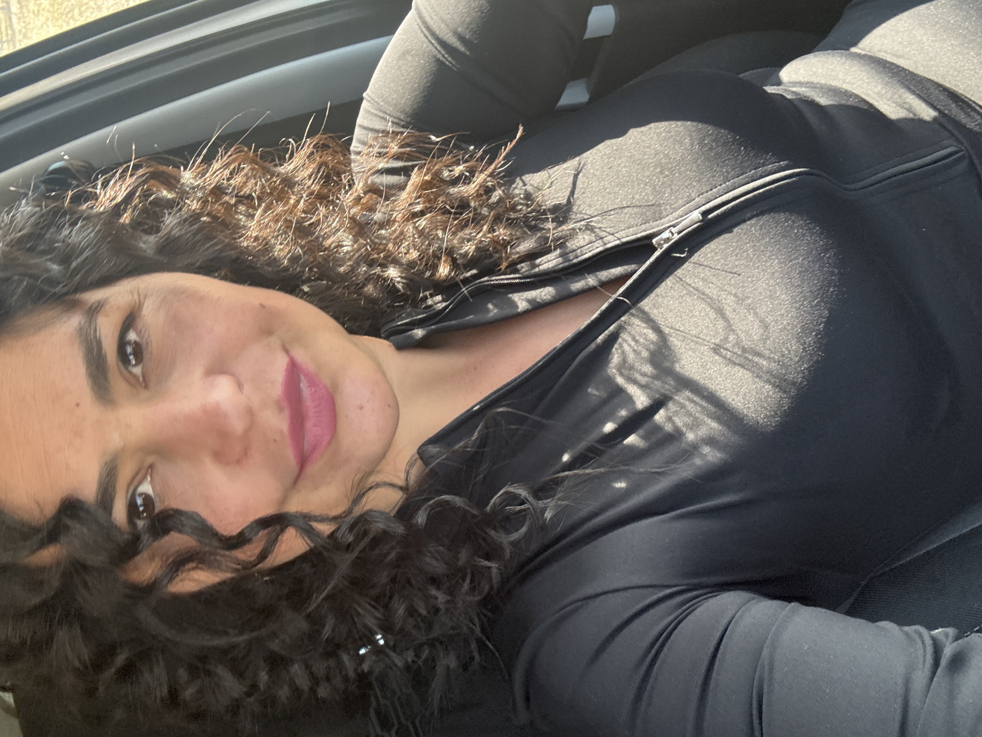 A close-up, eye-level portrait shows a woman inside what appears to be a car, likely a selfie. She is positioned horizontally, with her head resting or angled back, looking directly at the viewer with a gentle, inviting smile. Her dark, curly hair frames her face, with individual strands catching bright sunlight, particularly on the left side of the frame. She has warm brown eyes and is wearing pink lipstick.
She is dressed in a black, athletic-style top or jacket, featuring a prominent zipper down the front. The fabric appears to be textured, possibly a performance material. Strong, directional sunlight casts clear highlights and shadows across her clothing and hair, indicating a bright, sunny day. Part of a car window frame is visible in the upper left, reinforcing the indoor-vehicle setting. No text is visible in the image. The scene is set in Кемуль, Russia.