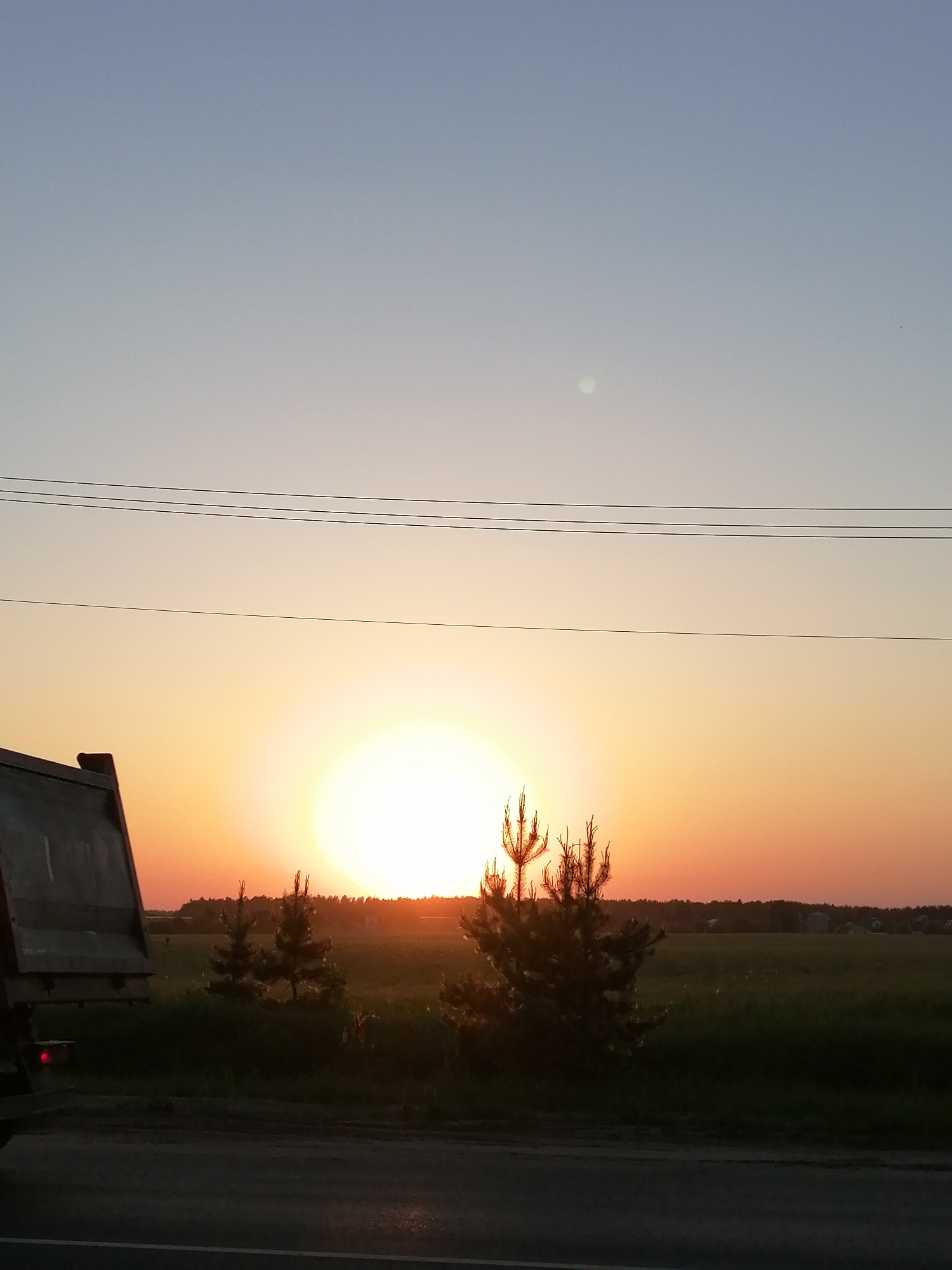A landscape photograph depicts a bright sunset over a field in an unspecified location, presumably a rural area. The sky transitions from a pale blue at the top to a vibrant orange and yellow as it nears the horizon. Three power lines run horizontally across the upper third of the image. The sun, a large, diffuse orb of light, dominates the center of the frame.
In the foreground, a road stretches across the bottom of the image, reflecting some of the sunset's light. To the left, part of a large truck, likely a dump truck, is visible, its bed dark against the bright sky. Three young pine trees are positioned between the truck and the field, their silhouettes adding depth to the scene. The field extends into the distance, with a tree line visible on the horizon. The environment suggests a peaceful, undeveloped area.