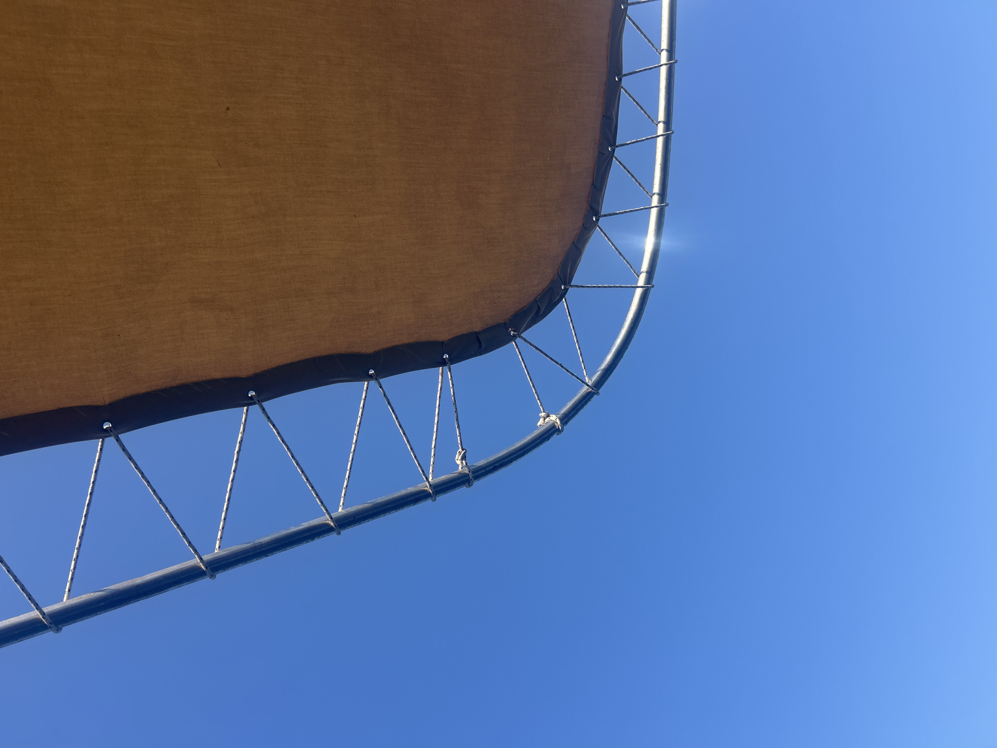 The image shows a close-up, worm's-eye view of a trampoline mat and its frame against a clear blue sky. The trampoline mat is a light brown fabric, stretched taut over a circular, silver-colored metal frame. The frame is supported by numerous metal springs, which are attached to the frame at regular intervals. The springs are connected to the edge of the mat by means of grommets and short ropes, creating a triangular pattern between the frame and the mat. The sky is a vibrant, cloudless blue, indicating a sunny day. The location is the city of Neo Chorio, Cyprus. There are no people or animals visible in the image.