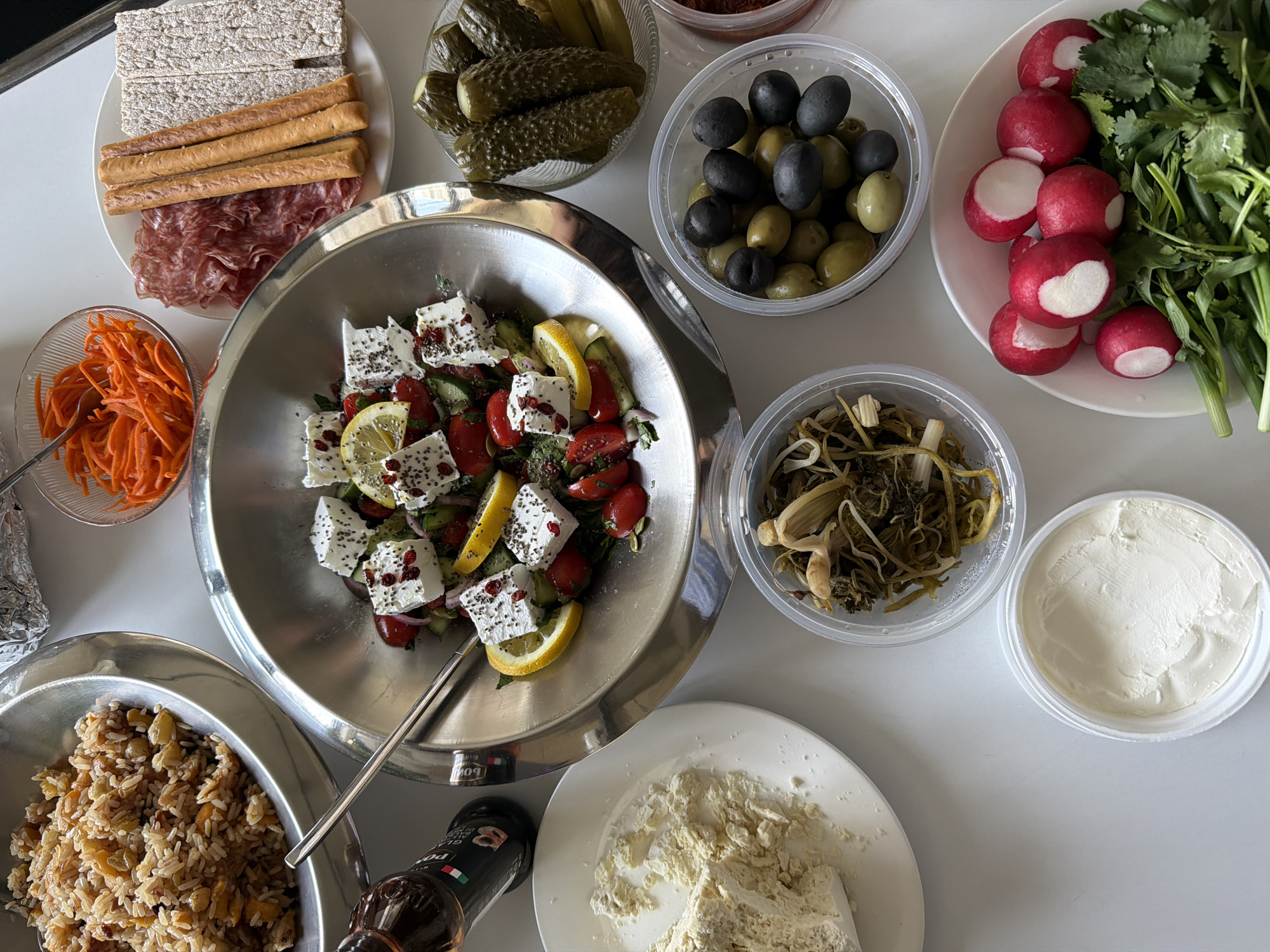 A high-angle shot depicts an assortment of food items arranged on a white surface. The arrangement suggests a meal preparation setting.
Food items include:
- A silver bowl filled with a salad comprised of feta cheese cubes, tomatoes, cucumbers, lemon slices, and seasonings.
- A plate containing crackers, breadsticks, and sliced meat (possibly salami or prosciutto).
- A bowl of pickled cucumbers.
- A container of mixed black and green olives.
- A plate of radishes alongside green herbs.
- A container of what appears to be pickled vegetables.
- A container filled with a white creamy substance (possibly a dip or cheese).
- A plate of what looks like crumbled cheese.
- A bowl containing a rice and vegetable dish.
- A bowl of shredded carrots.
- A bottle of dressing or condiment.
The scene does not contain any people.
There is no location in Yerevan, Armenia visible in this image.