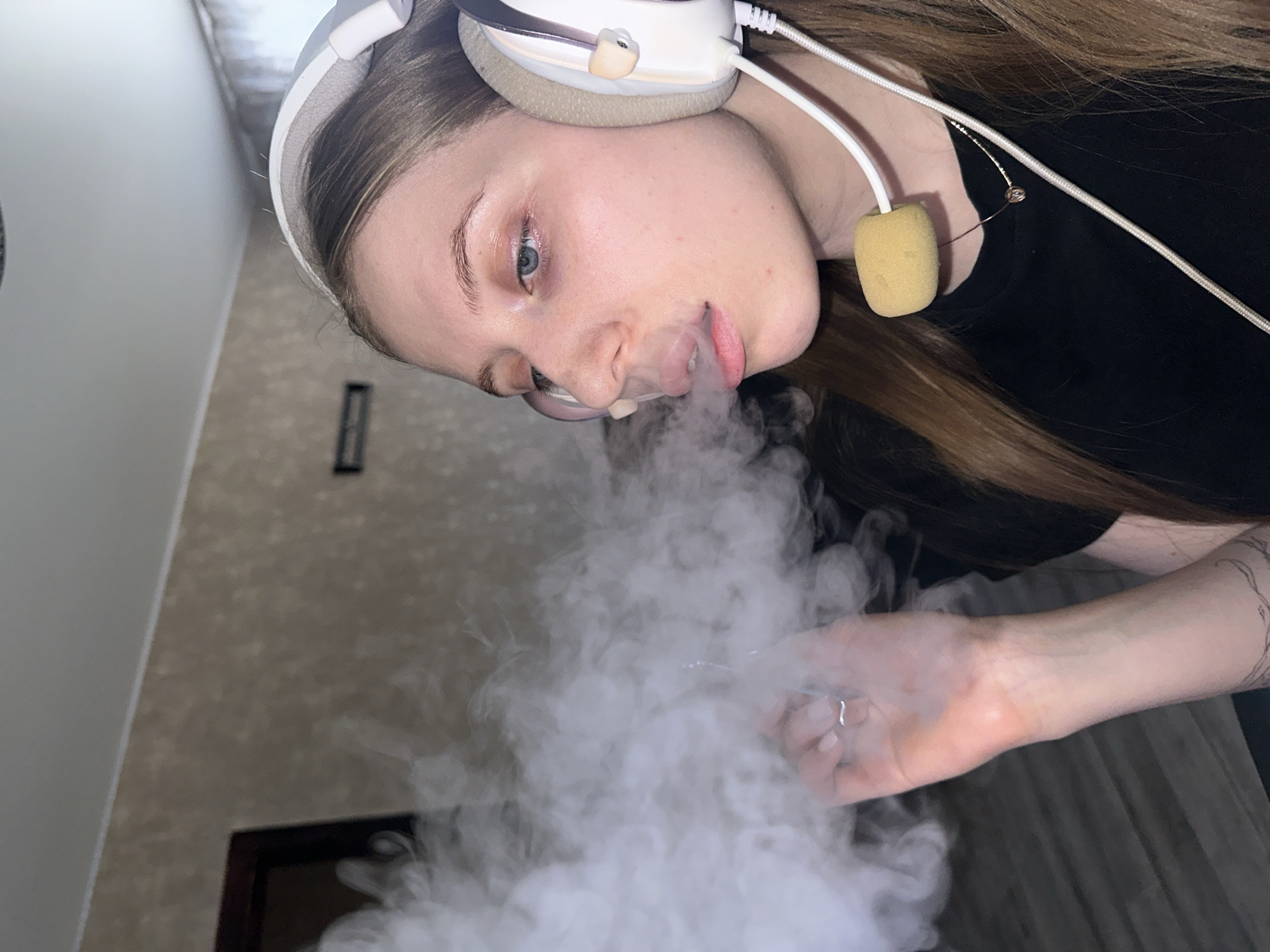 A young female, with light brown hair and blue eyes, is depicted in profile in an indoor environment. She wears white over-ear headphones with a yellow foam-covered microphone boom and a black top. A thin chain necklace with a small pendant is visible. Her left hand, adorned with a silver ring on the ring finger and a tattoo on the forearm, is positioned in the left foreground. She is exhaling a dense white vapor cloud from her mouth, obscuring the vaping device she is holding. The background consists of a light brown textured wall, a dark brown door frame on the left, and a white ceiling. A dark, horizontal object is mounted on the wall. The flooring appears to be dark wood or laminate. The specific location within the city of Chaikovsky, Russia, cannot be determined from the visual evidence.