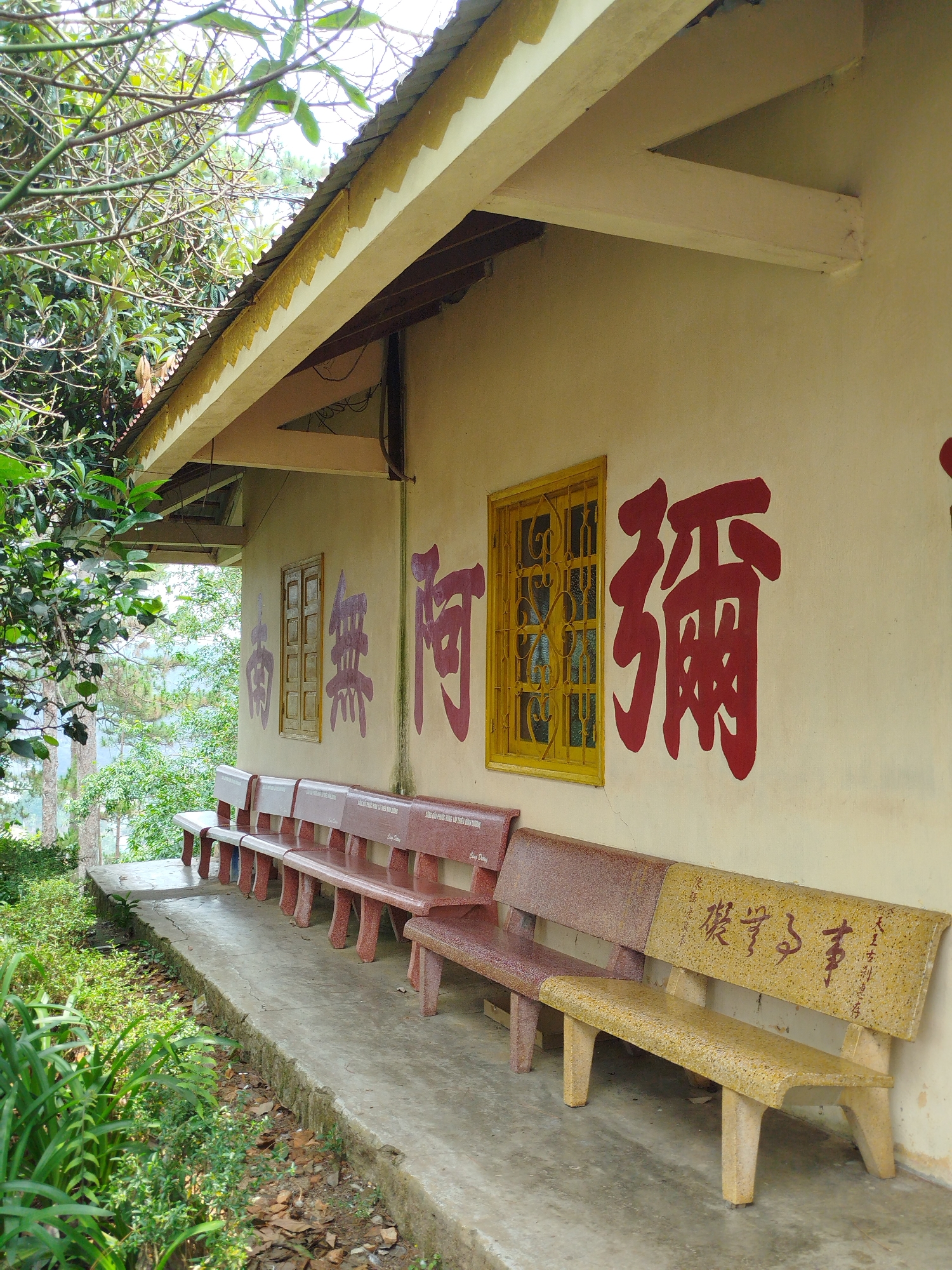 The image shows a row of stone benches in front of a building with Chinese characters painted on the wall. The benches are arranged on a concrete platform, and there is vegetation growing along the edge of the platform. The building has a cream-colored wall, a window with yellow bars, and a corrugated metal roof with a decorative edge. The Chinese characters are painted in red and purple. The location is likely a temple or other religious site in Da Lat, Vietnam. There are no people visible in the image. The environment is somewhat overgrown and natural.