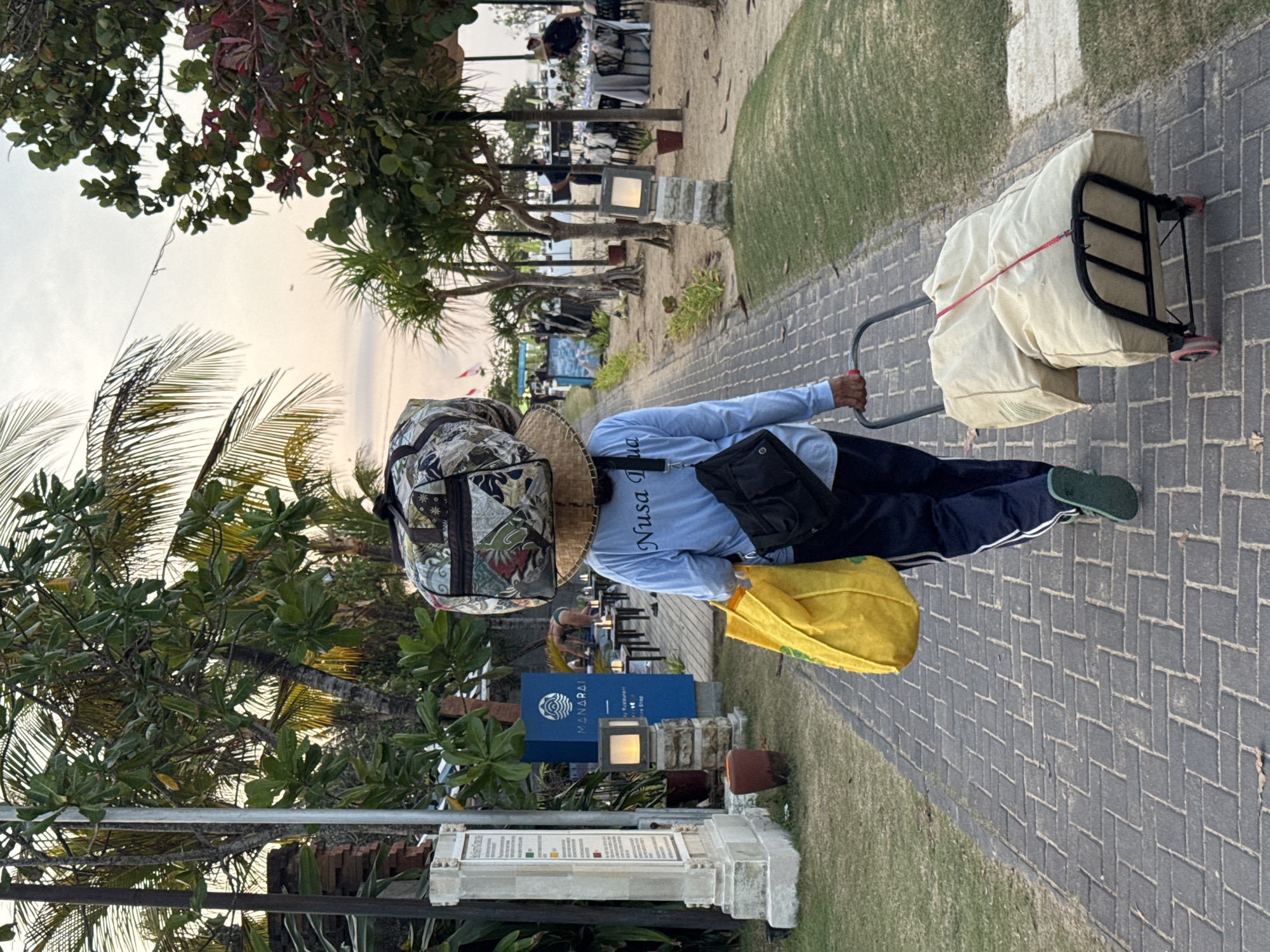 In Kuta Selatan, Indonesia, a person walks away from the camera on a brick pathway. They wear a blue long-sleeved shirt with "Nusa Dua" written on the back, dark pants with white stripes, and green flip-flops. On their head, they balance a wide-brimmed hat and a patterned duffel bag. A large yellow tote bag hangs from their left shoulder, and a black crossbody bag is on their right. They are pulling a black trolley cart laden with two large, beige-covered items secured by a red strap. To the left of the pathway, a stone monument displays information, and a blue sign with the word "MANARAI" is visible. Beyond that, a restaurant with tables and chairs is set up on a sandy area, with palm trees and other greenery surrounding the scene. The sky above is overcast.