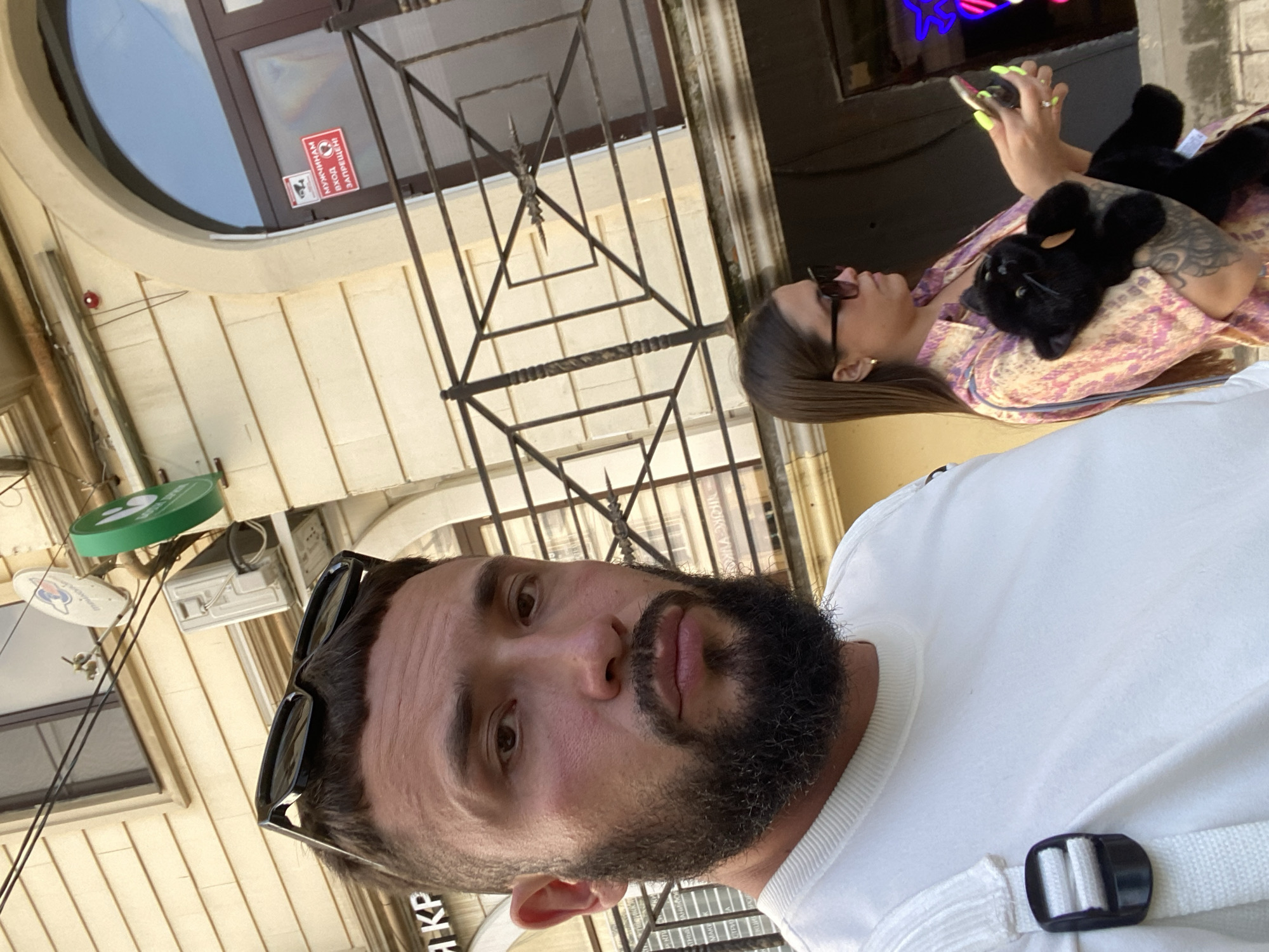 This is a selfie taken in Derbent, Russia, during the daytime. The photographer, a man with a beard and sunglasses, is in the foreground. In the background, a woman is holding a black plush toy and looking at her phone. She is wearing a patterned dress and sunglasses. Behind them, there is a building with a large, circular window and metal bars. There are also some signs visible. One red sign on the window has text in Cyrillic that appears to say "ОТКРЫТО" (OPEN) and some symbols. Another green sign is also visible, though the text is not clear. The overall setting appears to be an urban environment, possibly an outdoor cafe or street. The lighting suggests it's a sunny day.