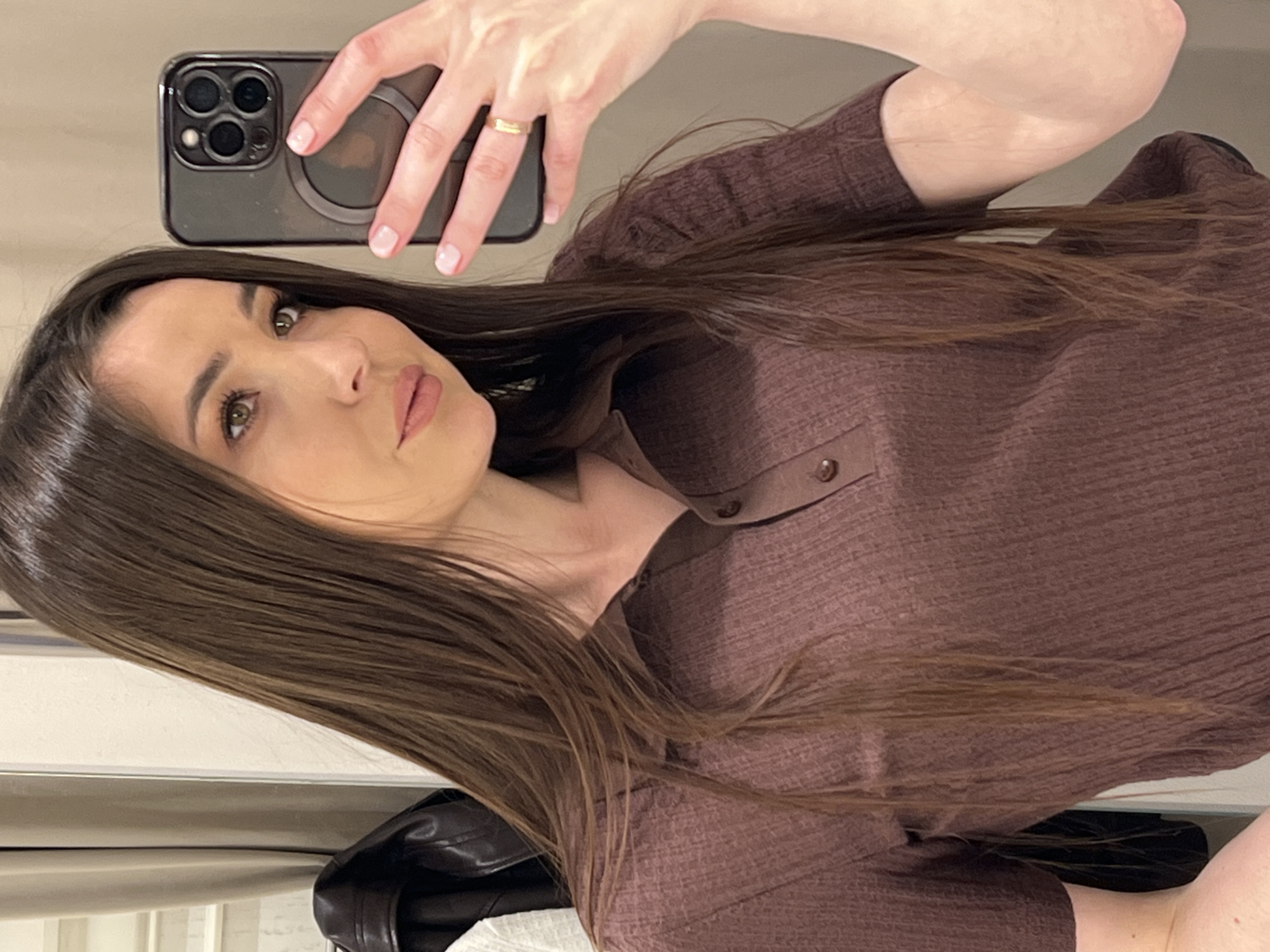 The image is a selfie of a woman, framed from the head to the upper torso, taken in an indoor setting in what is contextualized as Moscow, Russia. She has long, straight, dark brown hair that falls over her shoulders and chest. The woman is looking directly into the camera with a neutral expression, her eyes visible and her lips painted with a natural-toned lipstick. She appears to be wearing natural makeup.
She is holding a dark-colored smartphone, likely an iPhone with a triple camera array, which is encased in a clear cover featuring a circular MagSafe ring. Her left hand, with light pink manicured fingernails, grips the phone, and a simple gold-colored band is visible on her ring finger. She is dressed in a ribbed, collared shirt of a deep brown or maroon color, with buttons visible at the neckline. A dark, possibly leather, garment or jacket is partially visible in the bottom left corner of the frame. The background is a soft, light color, suggesting an interior wall or plain furniture. No specific time of day, weather, or visible text can be discerned from the image itself.