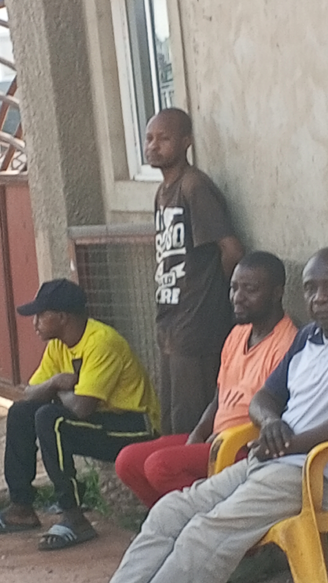 Four males are positioned outside of a building. One male stands against the building; he wears a black T-shirt with white text. A male sits on the ground to the left; he wears a black baseball cap, a yellow shirt, and black athletic pants with a yellow stripe. Two males sit in yellow plastic chairs to the right. One wears an orange shirt and red pants. The other wears a white shirt, a navy blue jacket, and gray pants. The location is Maiburiji, Nigeria.