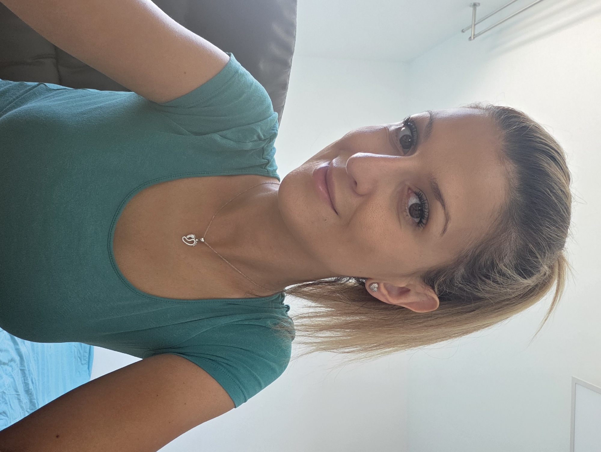 The image is a selfie of a woman with blonde hair pulled back in a ponytail. She is wearing a teal-colored t-shirt and a silver necklace with a heart-shaped pendant. She has on small heart-shaped earrings. The background is a plain white wall with some light fixtures. The woman appears to be indoors, likely in a home or office. No specific location in Munich, Germany can be determined from the image.