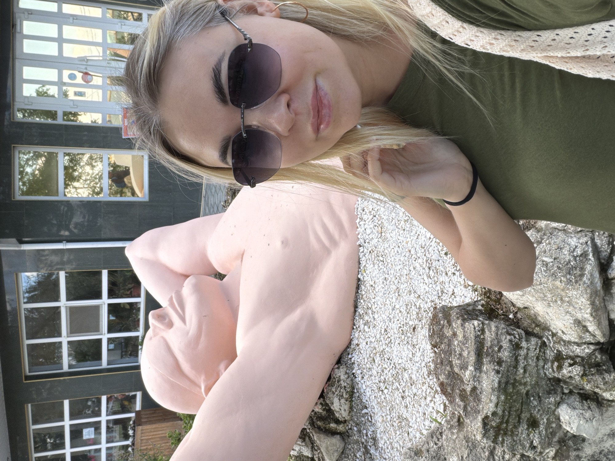 A woman is photographed outdoors in Anapa, Russia. She is wearing sunglasses, a green shirt, and a light-colored sweater. She holds her blond hair near her face. To her right is a large, reclining pink statue on a gravel surface near a building with windows. The setting appears to be a park or recreational area.