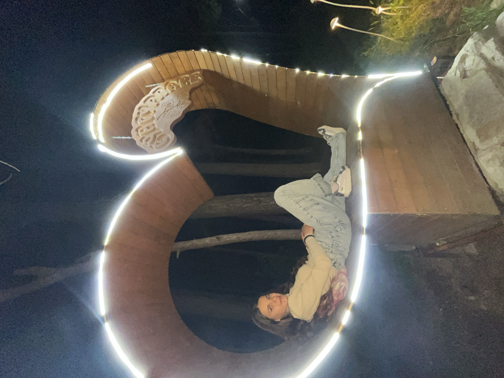 A nighttime, medium-shot photo depicts a young woman in jeans and a beige jacket, positioned within a large, illuminated, Cyrillic letter "Я". The structure appears to be made of wood, edged with bright white lights. The location is Sungurlare, Bulgaria, indicated by Cyrillic script incorporated into the letter's design. The woman is looking at the camera. The surrounding environment is dark.