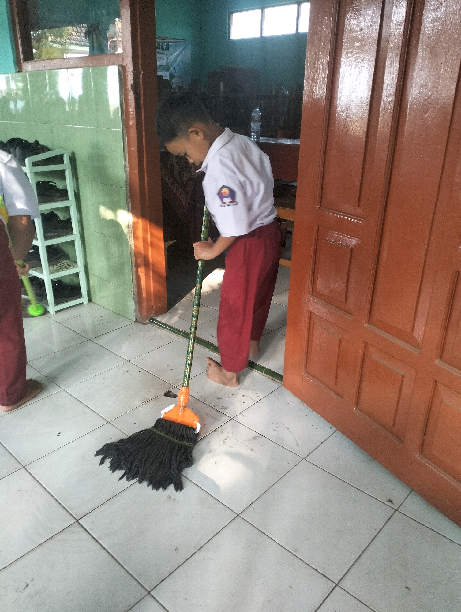A young boy, dressed in a white school shirt and red pants, sweeps a tiled floor with a mop. He is barefoot. The mop has a black head and an orange plastic connector attached to a green and black striped pole. To the left, another person in similar school attire, only their legs are visible, is also holding a green mop. The scene appears to be indoors, possibly a classroom or school hallway, with green tiled walls on the left and a large brown wooden door on the right. A shoe rack with several pairs of shoes is visible against the green wall. The floor tiles are a light color and appear slightly dirty. The background shows hints of furniture and windows. The overall impression is of a school cleaning activity in Blabakan, Indonesia.