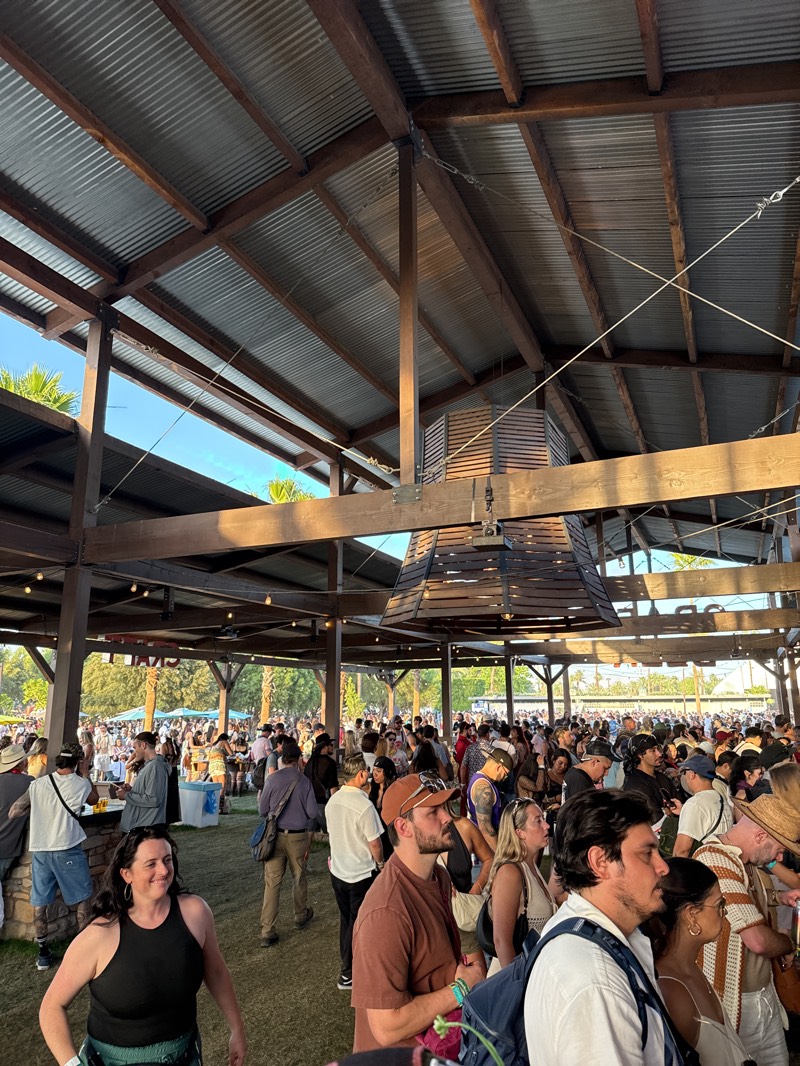 The image shows a large crowd of people gathered at an outdoor event in what appears to be a park or open space. The event is taking place under a large, open-sided structure with a corrugated metal roof supported by wooden beams. A distinctive, multi-tiered, slatted, wooden fixture hangs from the roof. Palm trees are visible in the background. The people in the crowd are diverse in appearance and are dressed in casual clothing. Based on the setting and the time of day, it is likely a concert or festival. Given the location of Indio, California, United States, it could be the Coachella Valley Music and Arts Festival or a similar event. The lighting suggests it's late afternoon or early evening.