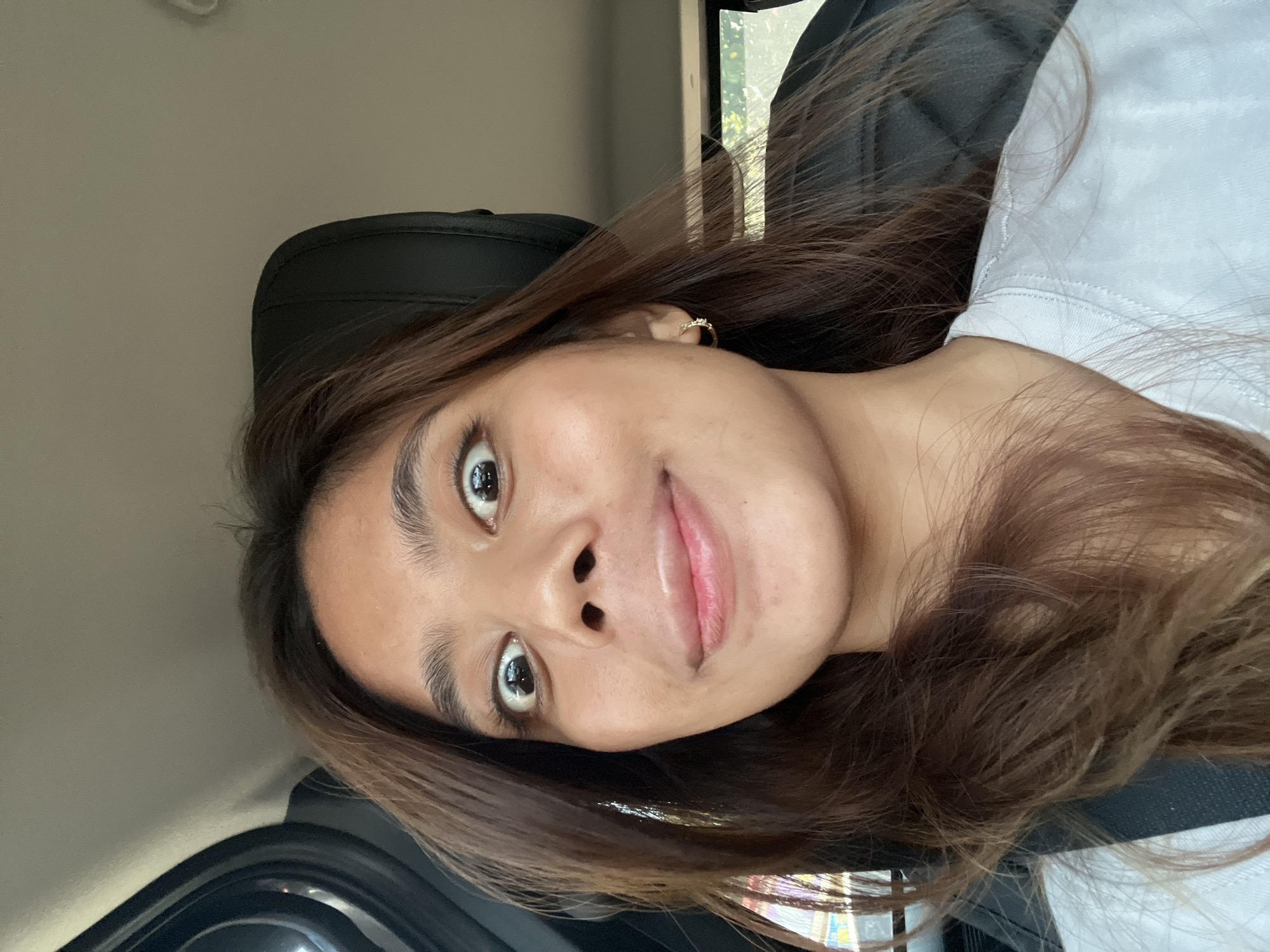 A young adult female with medium brown hair is depicted inside the passenger cabin of a vehicle, possibly in Davao City, Philippines, during daylight hours. She is looking directly at the camera with a wide-eyed, expressive gaze, her lips slightly parted in a faint smile or smirk. Her hair is parted roughly in the middle and falls over her shoulders, with some strands obscuring parts of her face. She is wearing a plain white short-sleeved t-shirt and a dark-colored seatbelt diagonally across her chest and shoulder. A small, gold-colored hoop earring with a minor embellishment is visible on her right earlobe. The vehicle's interior features a black textured seat headrest and a light-colored ceiling. Through the car windows, blurred reflections and background elements suggest an outdoor urban or suburban setting, including indistinct structures, some green foliage, and what appears to be part of a distant building or sign. No specific identifying landmarks of Davao City are discernible from the image alone, but the general street environment is consistent with a city location.