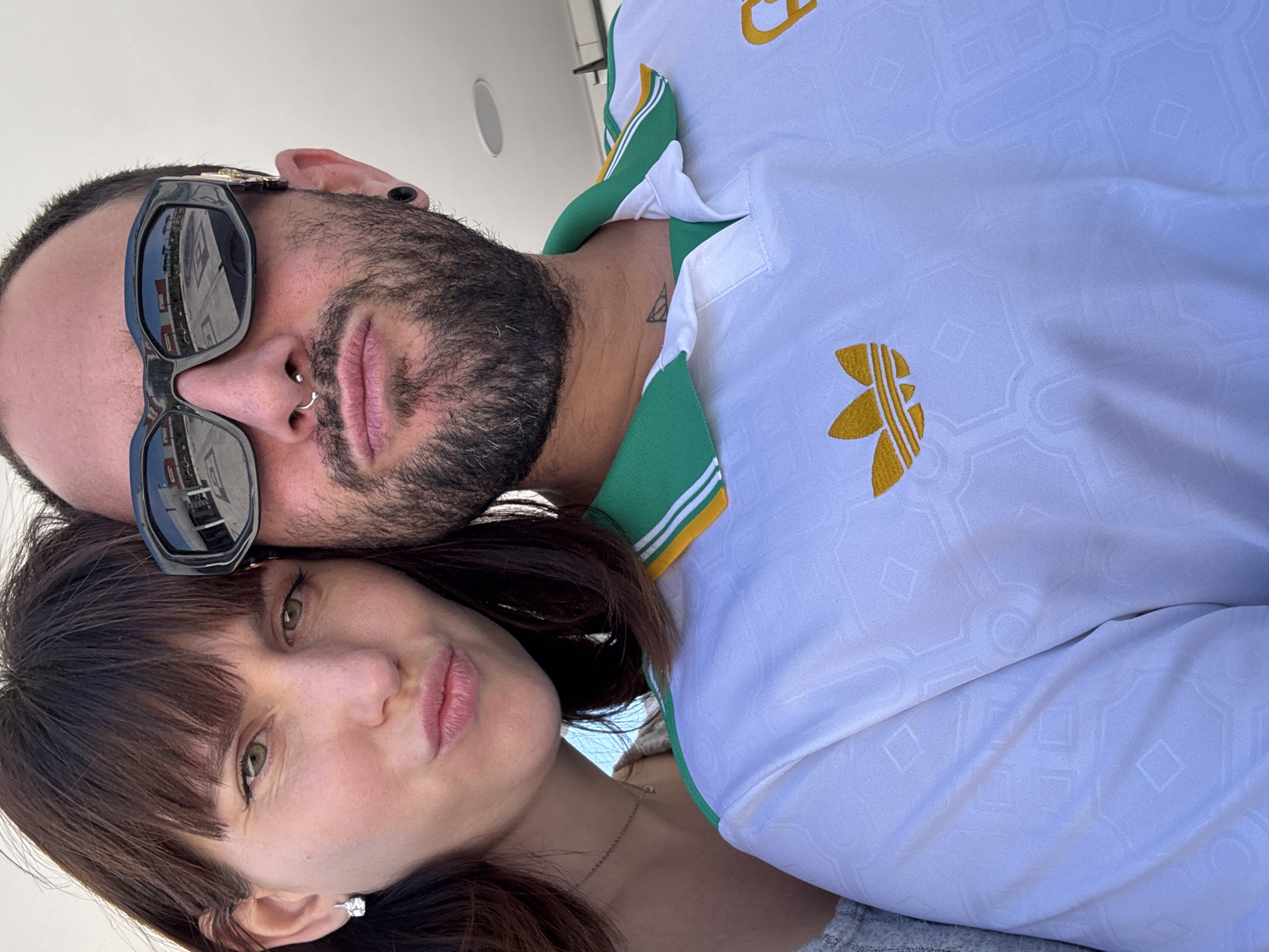 A selfie, taken at an upward angle, shows a man and a woman close together. The man, positioned above, has dark hair, a beard, and sunglasses reflecting a bright urban setting. He also has a nose ring and what appears to be a tattoo on his neck. The woman is below him, with dark hair and a necklace. The man is wearing a white Adidas shirt with green and yellow accents. The background suggests an outdoor location, possibly in Pomezia, Italy, based on reflections in the man's sunglasses.