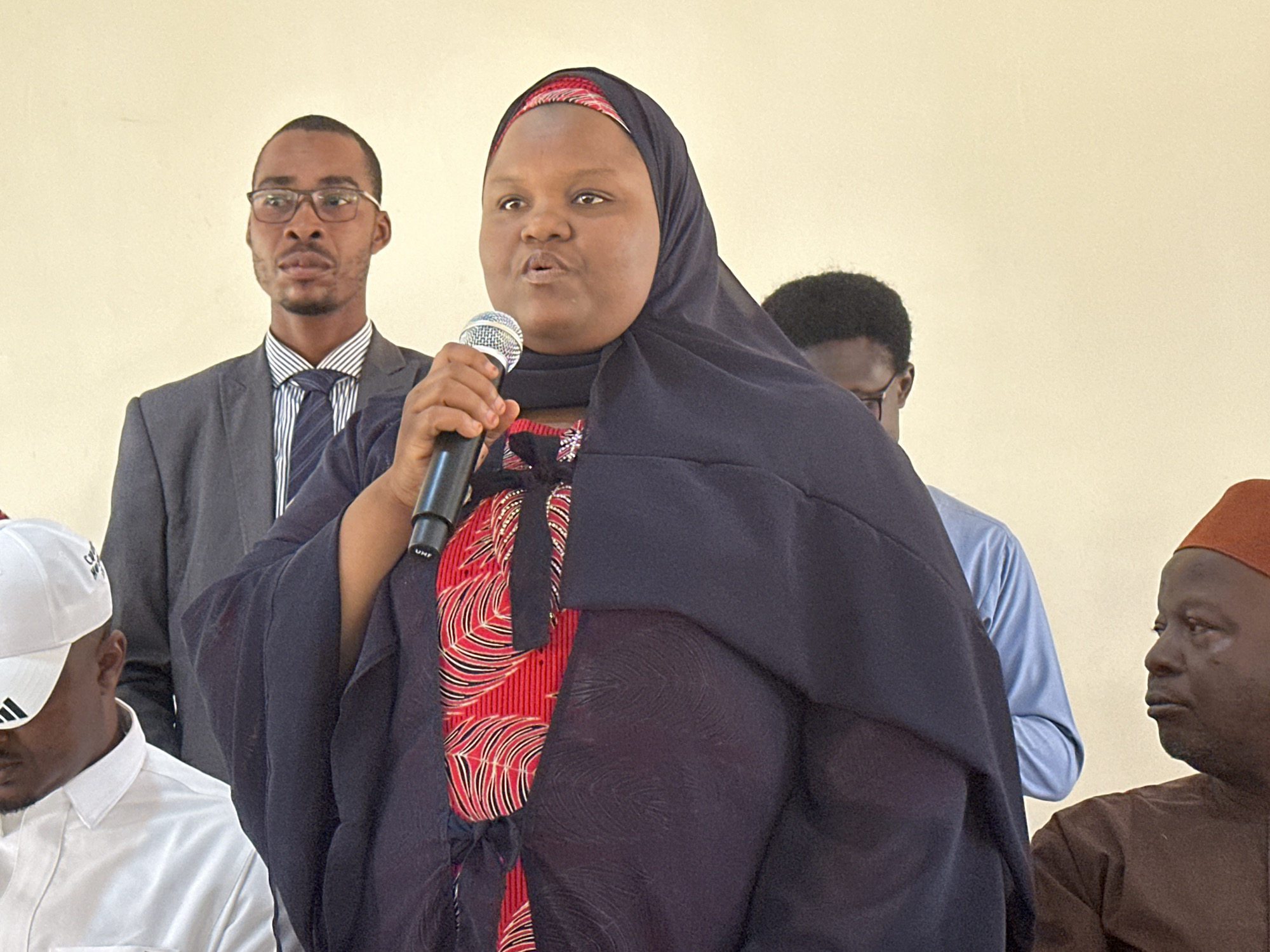 The image captures a woman speaking into a microphone. She is wearing a black hijab over a patterned red and black undergarment. A man with glasses and a suit stands behind her, and other people surround her in the foreground and background. The setting appears to be indoors with a plain wall behind the subjects. The location is listed as Unknown, Unknown.