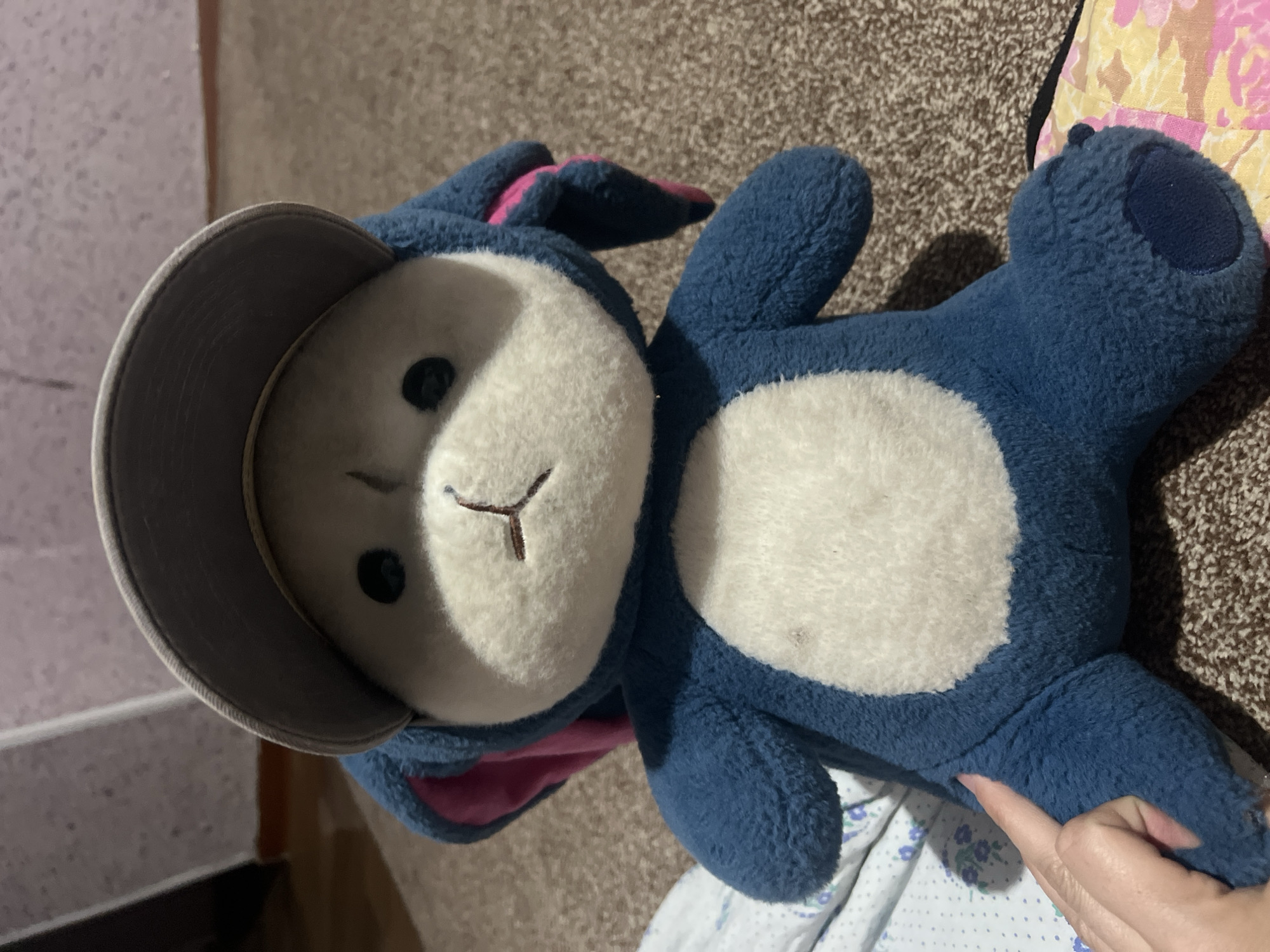 A blue plush bunny toy wearing a grey baseball cap is the subject of this close-up, eye-level shot. The toy is seated on a brown carpeted floor, with its left arm extended slightly and its right arm bent. A human hand with painted nails is visible in the lower left corner, gently touching the toy's left arm. The background is blurred, showing a wall with a pinkish-grey textured wallpaper and a wooden floor. The lighting is dim, creating shadows on the toy and the surrounding environment. The location is specified as Kara-Dıykan, Kyrgyzstan.