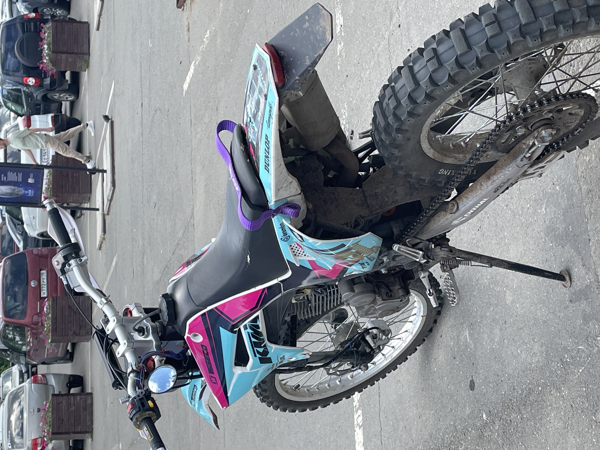 A dirt bike is parked on a paved surface, likely a parking lot, in what appears to be an urban environment. The dirt bike is the primary subject, with its front wheel turned slightly to the left. It features a vibrant color scheme with turquoise, pink, and black graphics. The bike is equipped with knobby tires, a chain drive, and a visible engine. A purple strap is attached to the seat. In the background, several cars are parked, including a red minivan with a license plate that appears to be Russian, and other vehicles that are less distinct. A man in a light green t-shirt and khaki pants is walking in the background, and several pigeons are on the ground. Wooden planters with flowers are also visible in the background. The overall scene suggests a casual urban setting.