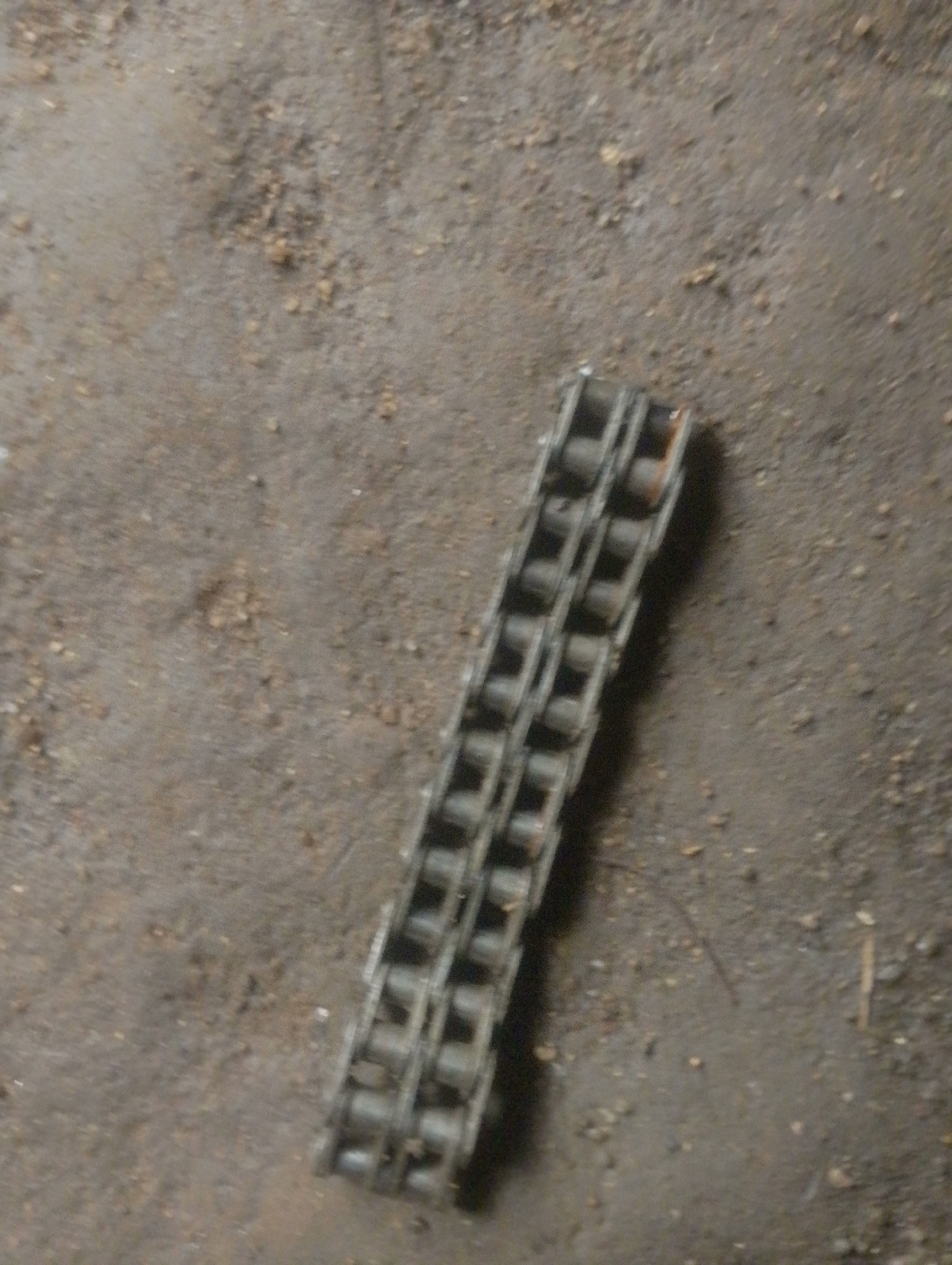 A close-up, overhead shot shows a section of a metal chain lying on a dusty, concrete-like surface. The chain is made up of interlocking links, each with rollers. The surface appears to be a floor or ground, with small pebbles and dirt scattered across it. There are no people or any other objects visible in the frame. The lighting is dim and seems to come from above, casting a slight shadow from the chain onto the surface. The scene is devoid of any specific geographical markers that would indicate its location within Jos, Nigeria, or any other city.