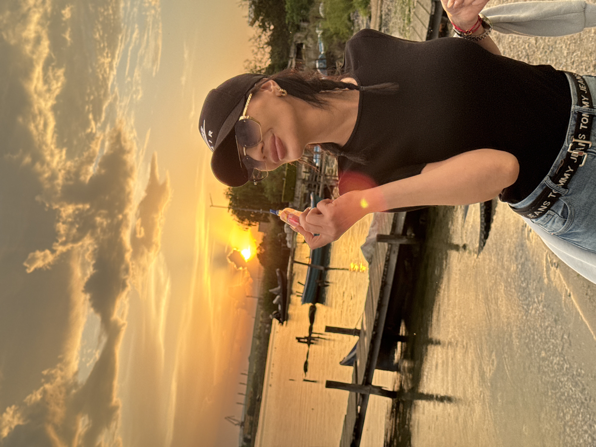 A woman stands on a dock in Казашко, Bulgaria, holding a snack, as the sun sets over the water. The sky is filled with golden clouds. The woman is wearing a black t-shirt, jeans with a Tommy Jeans belt, sunglasses, and a baseball cap. She is looking down at the snack in her hand. The dock is made of wood and extends out into the water. There are boats docked nearby.