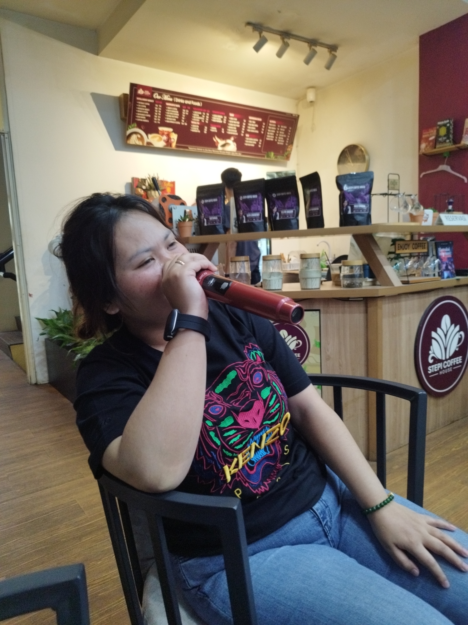 A young woman with dark hair is sitting in a black chair in what appears to be a coffee shop. She is wearing a black t-shirt with a colorful tiger graphic and blue jeans. She is holding a red object to her mouth, possibly a microphone or a vape pen. A smartwatch is visible on her left wrist. In the background, there are shelves with coffee bags, jars, and glasses, as well as a menu board on the wall. A circular logo for "Stepi Coffee House" is also visible on the counter. The environment suggests a casual indoor setting, likely a commercial establishment in South Jakarta, Indonesia.