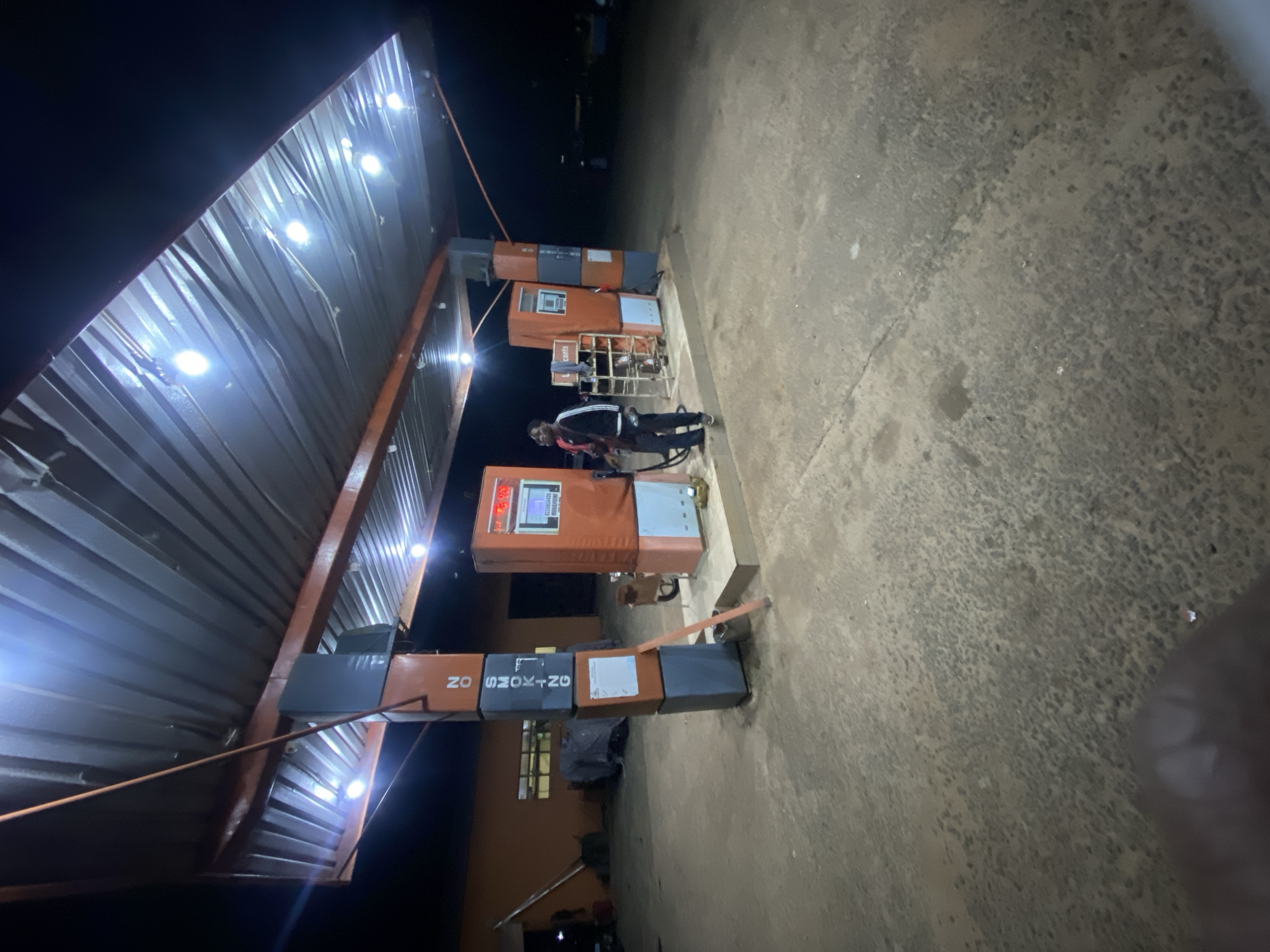 The image depicts a nighttime scene at a gas station in Sasaro, Nigeria. A male individual, clad in a dark jacket with white arm stripes and dark trousers, stands on a raised concrete platform beside an orange and white fuel pump. He is actively holding a fuel nozzle, appearing to dispense fuel. This pump displays red digital numbers and a screen below, where text including '8229950428' and 'BOSS PHONE' is discernible. A second, identical fuel pump is positioned to the left, partially obscured, with a 'Lubricants' label visible.
An orange-brown metal framework supports a corrugated metal canopy overhead, from which multiple bright white lights illuminate the station. Centrally located, a tall pillar is constructed from alternating orange-brown and dark gray rectangular blocks. The dark gray sections feature vertical white text stating 'NO SMOKING'. A small sign is affixed to an orange block, and a slender orange-brown rod leans against the pillar's base.
In the background to the left, a building with an orange-brown facade and gridded windows is visible, with a long pole or ladder leaning against it and tarp-covered items nearby. The ground is a textured concrete surface with visible cracks and variations in color. The areas beyond the station's direct illumination are dark.