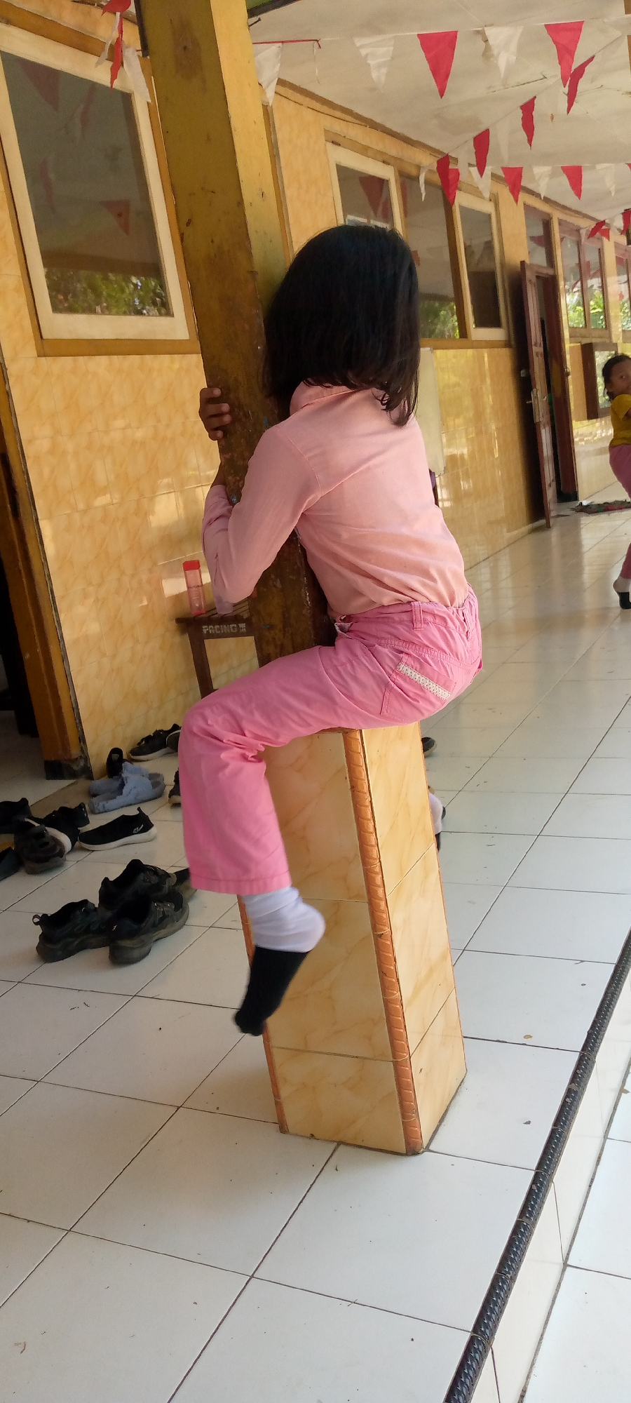 A young girl in a pink shirt and pink pants is perched on a wooden beam in what appears to be a covered outdoor walkway or school hallway. She is holding onto the beam with her arms and is sitting on a checkered, tiled floor. Her left leg is bent and her foot, wearing a black sock and a white sock over it, is extended. A small, red bottle and a small wooden stool are visible behind her, leaning against the beam. In the background, there are windows with yellow tiled walls and red and white triangular flags strung across the ceiling. Several pairs of shoes are scattered on the floor in front of the girl. Further down the walkway, another child in a yellow shirt and pink pants can be seen walking. The scene suggests a location in Pacing, Indonesia, possibly a school or community center.