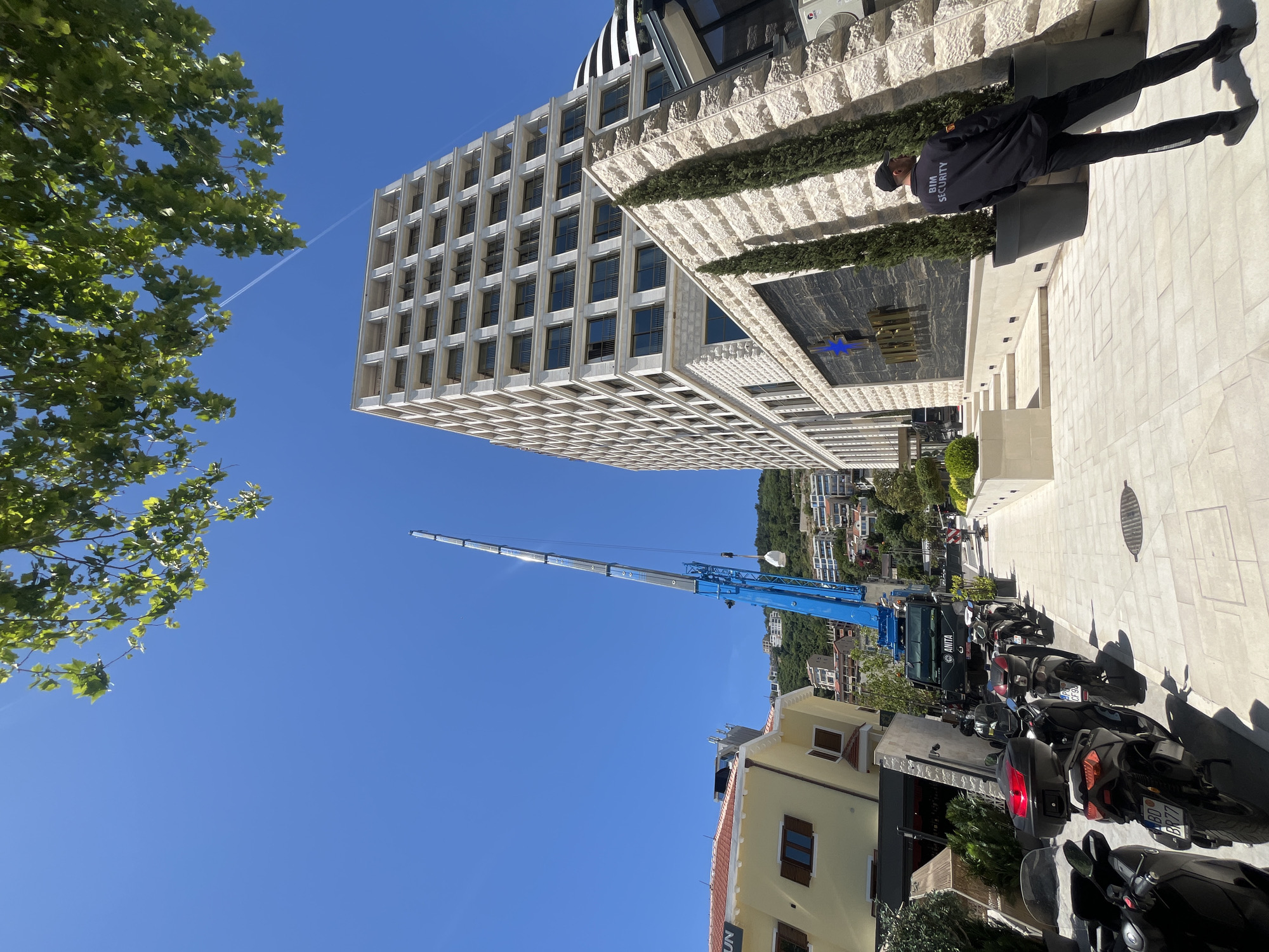 A street scene in Budva, Montenegro, features a multi-story building with a light-colored, patterned facade. A blue crane with an extended boom is parked nearby. A security guard wearing a black uniform stands in front of the building. Parked motorcycles line the street. The sky is clear and blue. Buildings and greenery are visible in the background, suggesting a residential area on a hill. A contrail is visible in the sky.