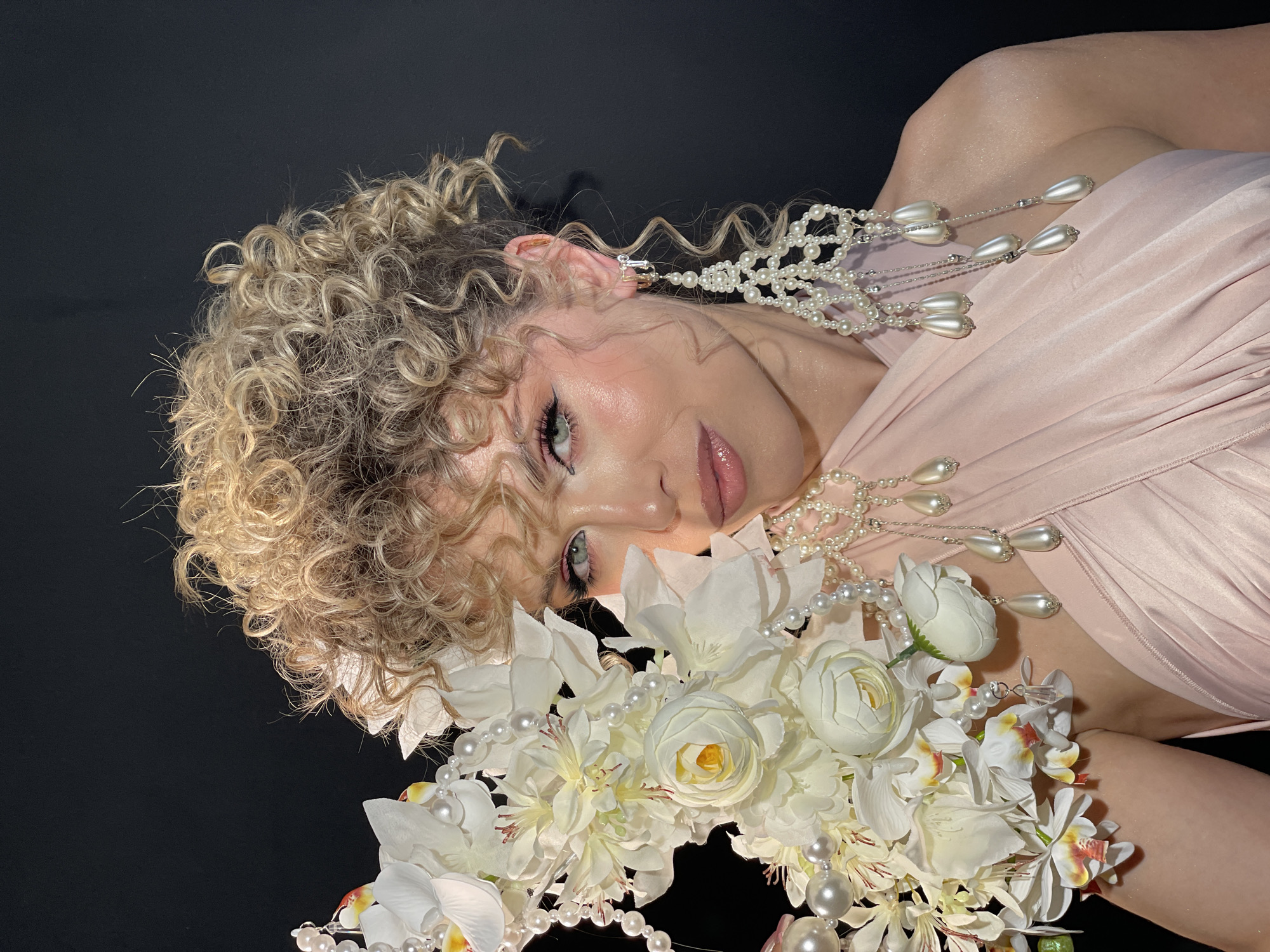 A woman with curly blonde hair is lying against a black background. She is wearing a light pink dress and pearl earrings. She is holding a bouquet of white flowers. The location is not discernible as Kazan, Russia.