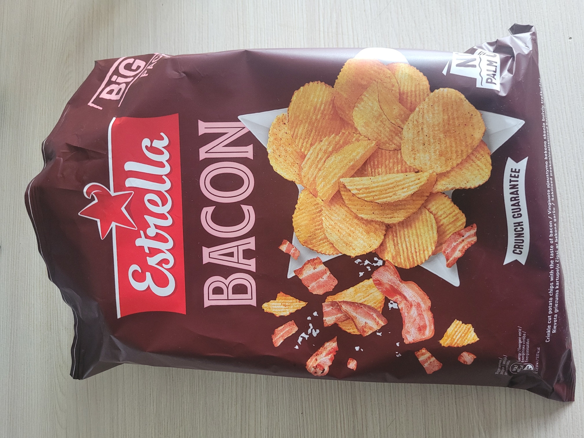 A bag of Estrella Bacon flavored potato chips is shown lying flat on a light-colored, textured surface, resembling a wooden table or countertop. The bag is primarily a deep maroon color with a prominent "Estrella" logo in white script on a red banner at the top. Below the logo, the word "BACON" is printed in large, pink, stylized letters. The bag features an illustration of crinkle-cut potato chips scattered with pieces of bacon and what appears to be sea salt. In the upper right corner, the word "BIG" is printed in white. Towards the bottom of the bag, there is text in various languages, including nutritional information, and a slogan that reads "CRUNCH GUARANTEE." A small logo indicating "NO PALM OIL" is visible on the lower right side of the bag. The lighting suggests an indoor environment, with a slight sheen on the plastic bag. There is no indication of the city of Jūrmala, Latvia, in the image.