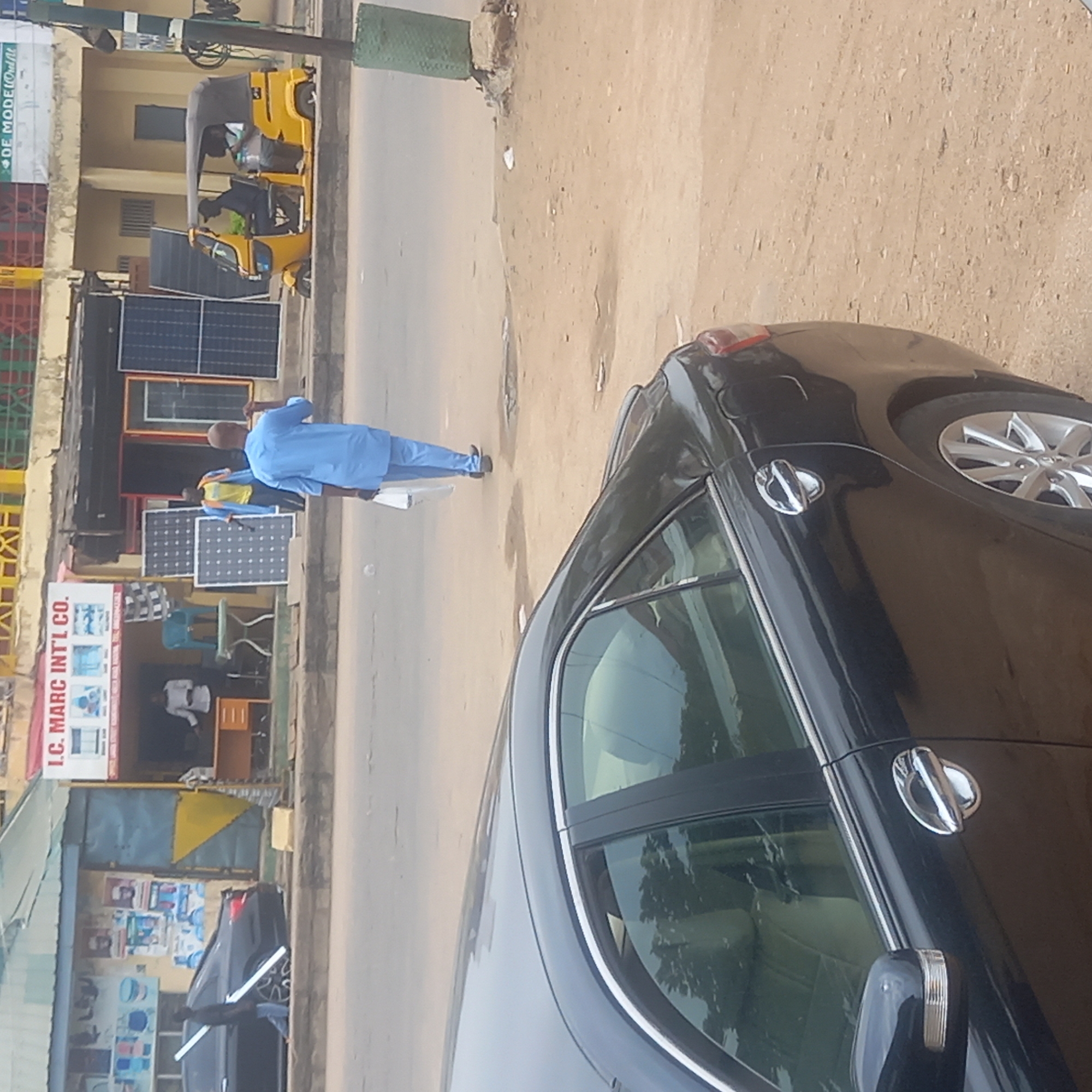 The image captures an urban scene in Dadi Riba, Nigeria. In the foreground, a portion of a black car is visible, including a side mirror, door handles, and a front tire. In the mid-ground, a man in a blue outfit is walking away from the camera on the road adjacent to a storefront. The storefront displays a sign reading "I.C. MARC INT'L CO.", with solar panels displayed nearby. Other people are visible inside the shop. In the background, there's a yellow three-wheeled vehicle (tuk-tuk) and additional buildings, giving a general sense of the urban environment.