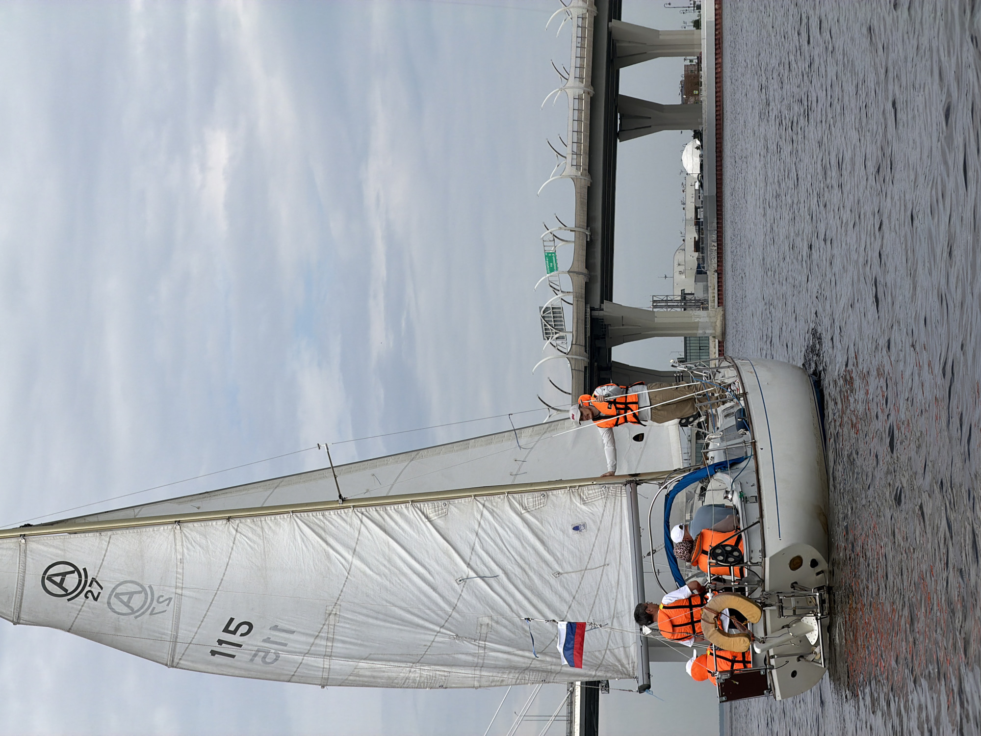 A white sailboat with a full mainsail sails on dark water under a cloudy sky. The mainsail displays the number "115" and some other markings. Several people wearing orange life vests are on board the sailboat. A Russian flag flies on the sailboat's mast. In the background, a large modern bridge with multiple lanes and concrete supports spans the water. The bridge appears to be part of the Western High-Speed Diameter in Saint Petersburg, Russia, given its characteristic design and the surrounding urban landscape which includes industrial buildings and a white domed structure. The scene depicts a recreational sailing activity near a major transportation infrastructure.