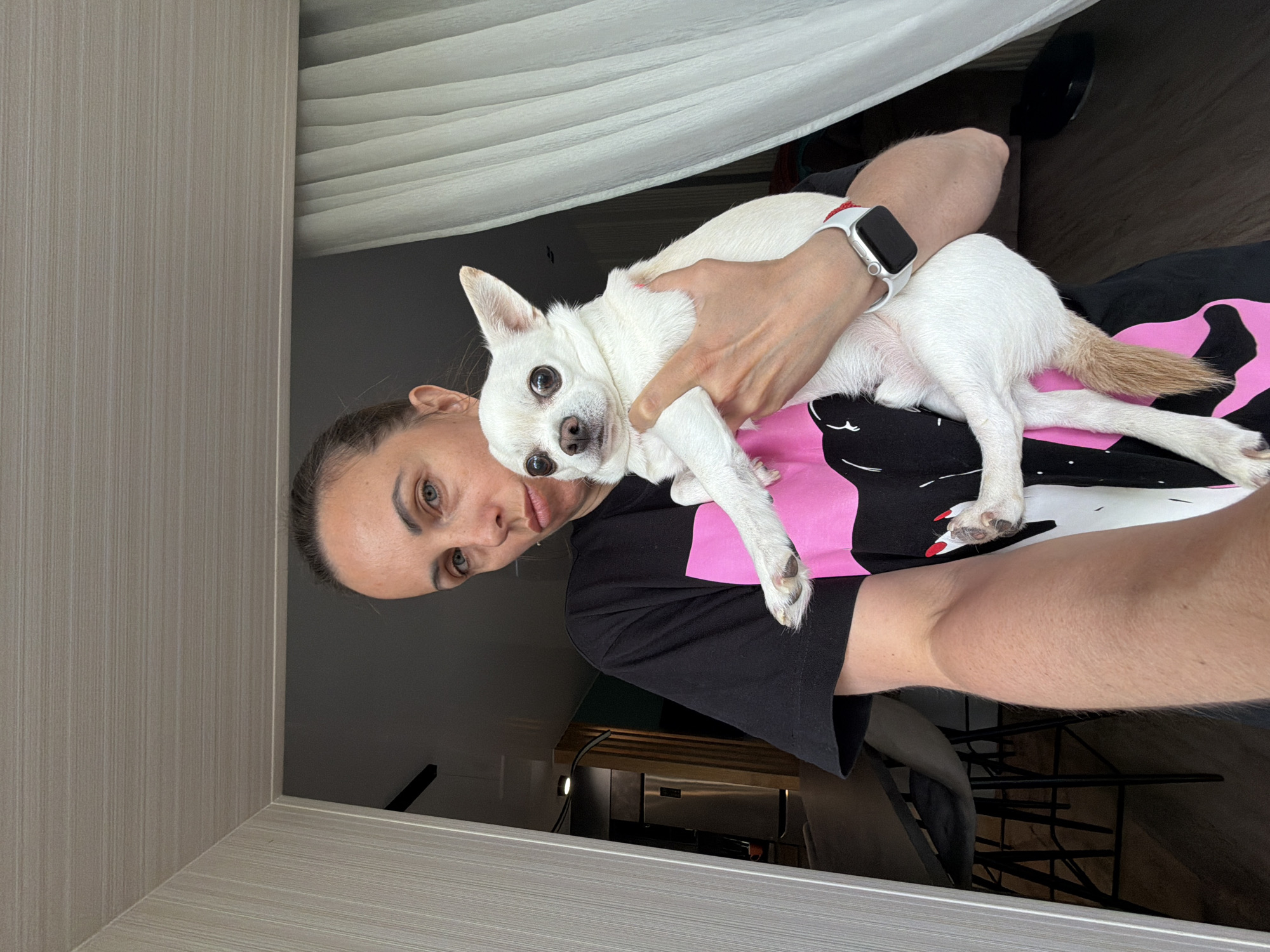A woman is holding a small white dog. The woman has light skin and dark hair pulled back from her face. She is wearing a black t-shirt with a pink and white graphic. The dog is primarily white with a tan tail. The woman is wearing a white smartwatch. The background includes part of a white curtain or drape, and what appears to be a room interior with furniture and a light-colored wall or paneling. There is no indication of the location of the image in Birobidzhan, Russia.