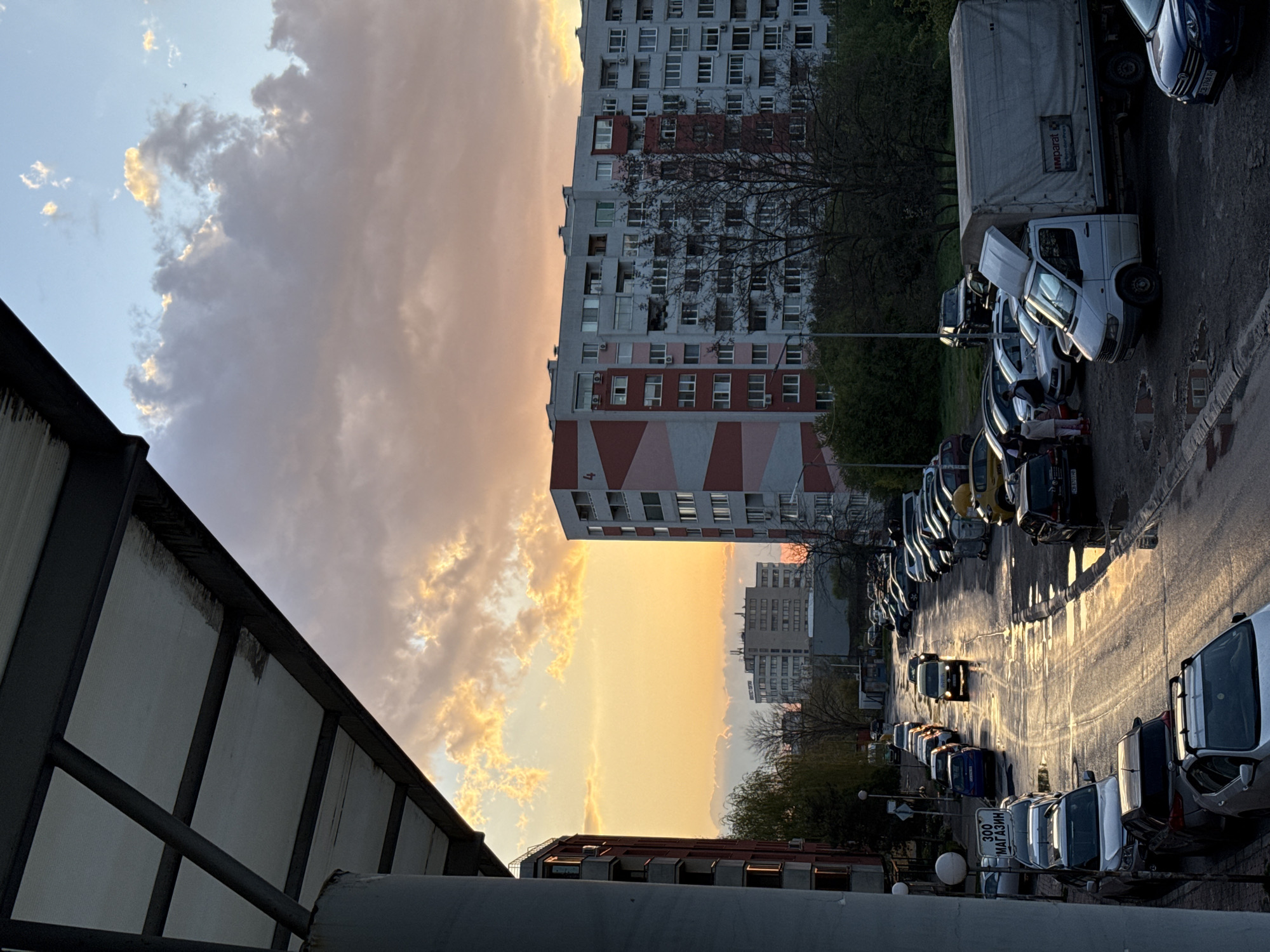 The image captures a street in Sofia, Bulgaria, at sunset. Tall apartment buildings dominate the background, illuminated by the golden light. Below, a street lined with parked cars reflects the sunlight. A pedestrian bridge is visible in the foreground, partially obscuring the lower portion of the scene. The street appears wet, potentially from recent rain. The sky is filled with clouds, some tinted orange by the setting sun.