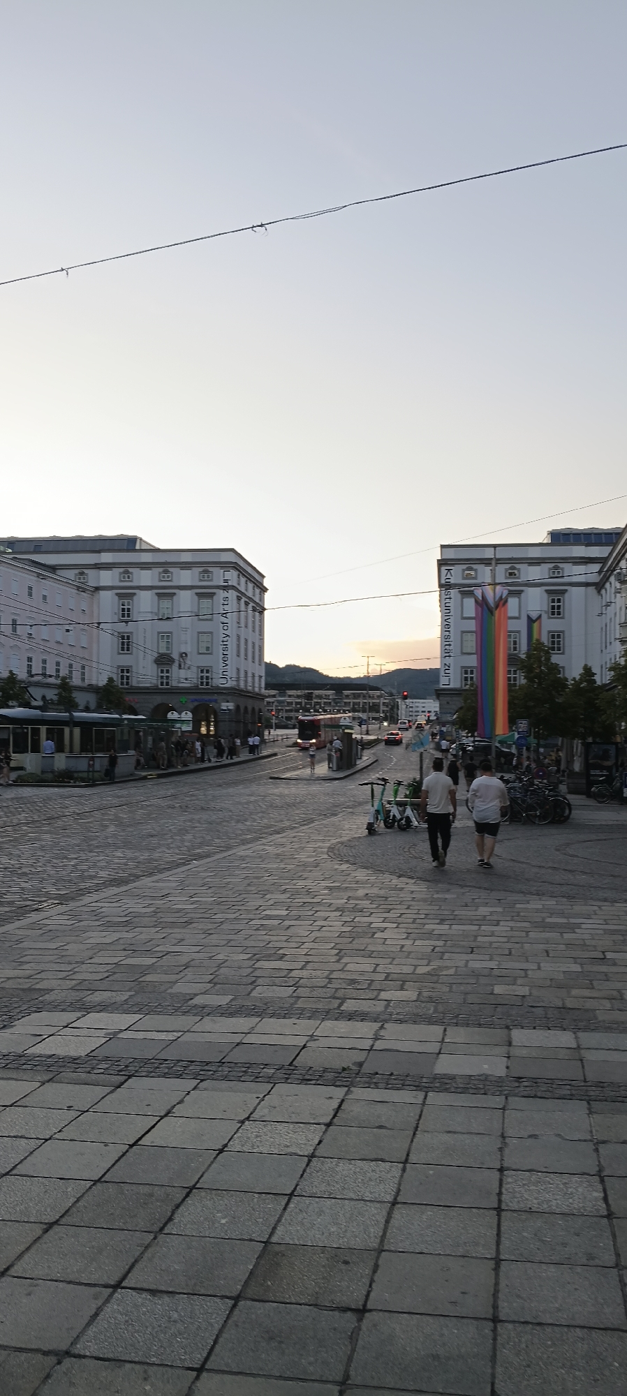 This is a vertical, eye-level shot of a cobblestone street in Linz, Austria, in what appears to be late afternoon or early evening. Two large, light-colored buildings flank the street, one on the left and one on the right. The building on the left has a banner that reads "University of Arts Linz." The building on the right also has a banner with the same text, along with a large rainbow flag hanging vertically beside it. The sky is pale and clear, with a hint of sunset colors in the distance.
The street is paved with cobblestones, and there is a tram track running down the center. A tram is stopped on the left side of the street, and several people are waiting at a platform. Further down the street, traffic is moving, with cars and a red bus visible. On the right side of the street, there are several parked electric scooters and bicycles. Two men are walking away from the camera down the street. In the background, there are trees and hills.