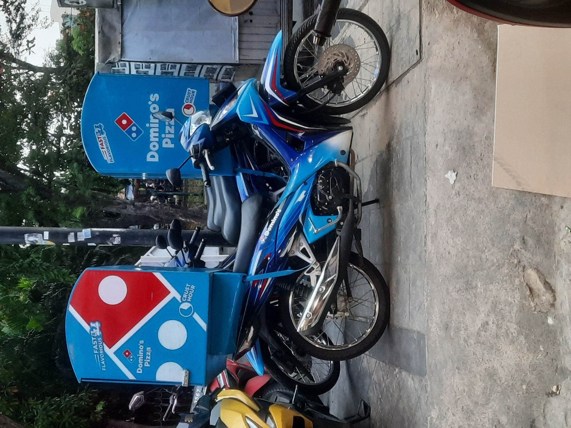 A blue Domino's Pizza delivery motorcycle is parked on a street in North Jakarta, Indonesia. The motorcycle is decorated with Domino's branding, including the company logo and slogans like "FAST & FLAVOROUS" and "CRUST HOUR." The motorcycle is equipped with a large blue delivery box on the rear, also branded with Domino's. The vehicle is parked alongside other motorcycles and is situated on a paved surface with some foliage in the background. The overall scene suggests a commercial or service-oriented environment within the urban setting of North Jakarta.