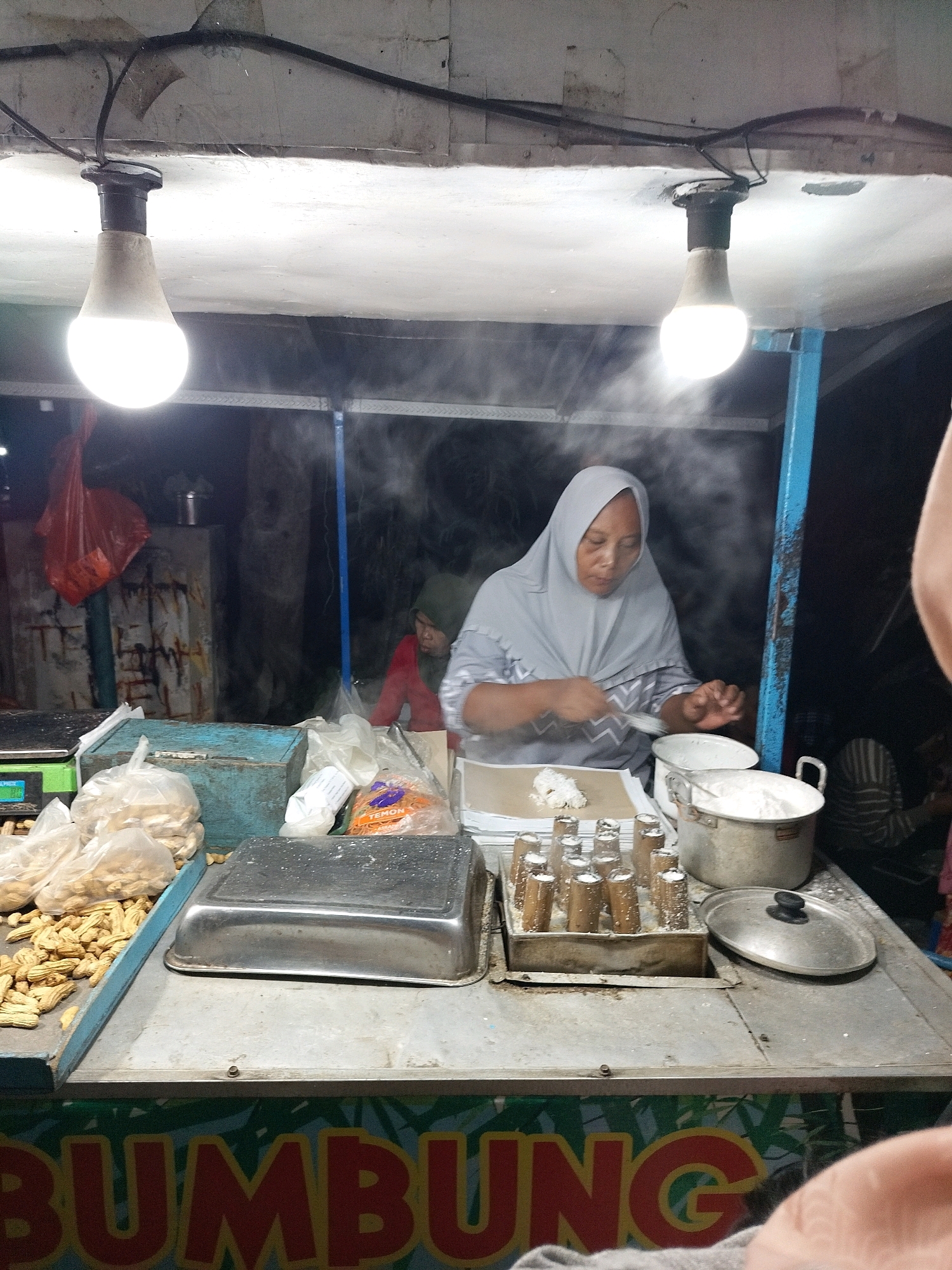 A street food vendor, wearing a grey hijab and grey patterned tunic, prepares food at a stall in Putat, Indonesia. The vendor is captured in the act of scooping or mixing ingredients on a paper-lined surface. In front of the vendor are several cooking pots, one filled with a white powder, and a tray containing numerous cylindrical molds filled with a white substance. To the left of the vendor, a scale is visible, along with bags of what appear to be peanuts. The stall is illuminated by two bright, hanging lightbulbs. Behind the vendor, a second person in a red shirt and dark head covering is partially visible. The stall has a metal countertop, and the background suggests a dark, outdoor environment, possibly at night. The word "BUMBUNG" is prominently displayed in large, red lettering on the front of the stall.