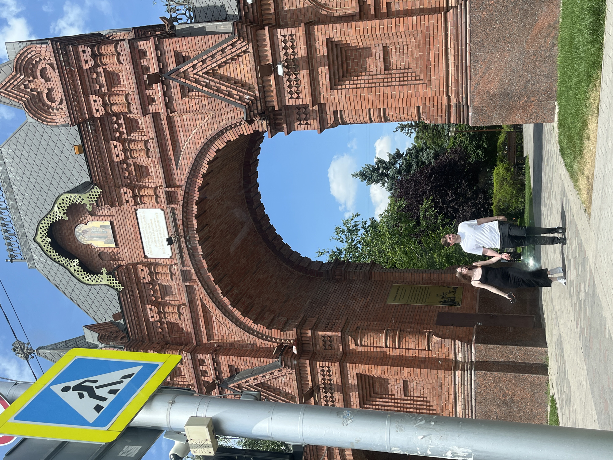 In an outdoor, daytime scene in Krasnodar, Russia, a young couple walks hand-in-hand on a paved walkway in front of a large, ornate red brick archway. The man on the right wears a white t-shirt and black pants, while the woman on the left wears a long black dress and white sneakers. They are walking towards the left of the frame. To the left of the couple, a pedestrian crossing sign with a blue background and white pedestrian symbol is prominently displayed on a metal pole, along with a traffic light and a control box. The archway, decorated with intricate brickwork and a dark grey roof, features an inscription and an image on its facade. Beyond the arch, lush green trees and a bright blue sky with scattered clouds are visible. The ground is a mix of paved surfaces and green grass.