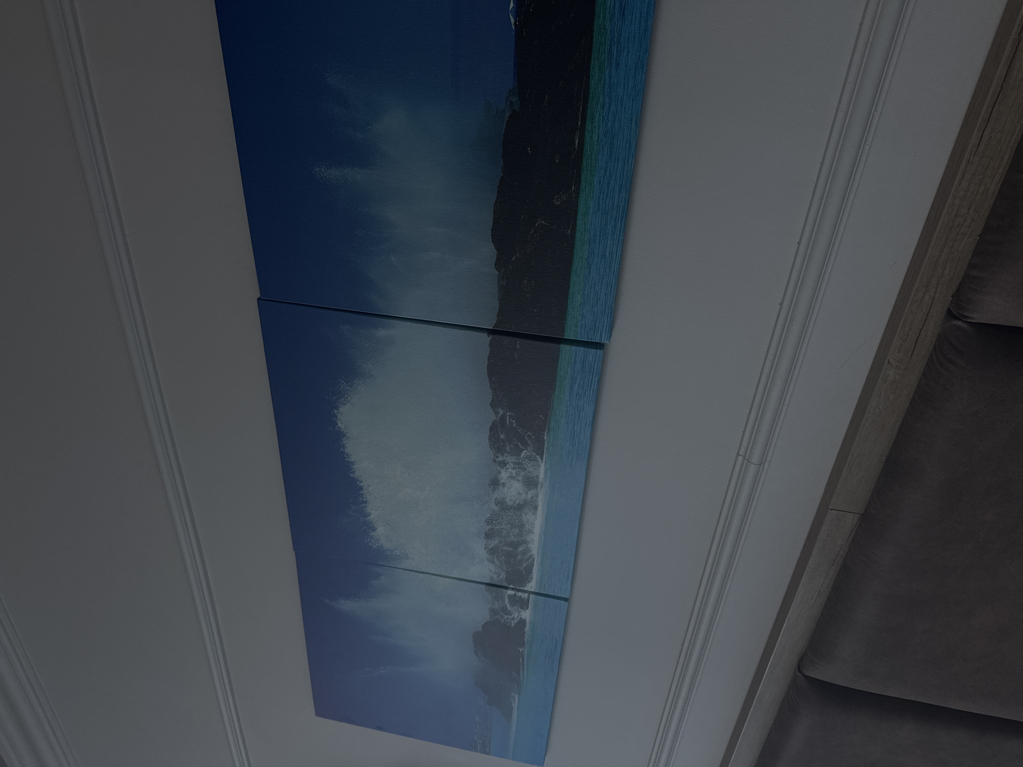 The image shows a three-panel canvas art piece hanging on a light gray wall. The artwork depicts a dramatic ocean scene with large waves crashing against dark rocks. The water is a vibrant turquoise, and the sky is a deep blue with white spray from the waves. Below the artwork, a portion of a dark gray upholstered headboard with a light-colored wooden trim is visible. The wall features ornate white crown molding along the top edge. The overall scene appears to be an interior shot, possibly in a bedroom or living room, in the city of Nesebar, Bulgaria.